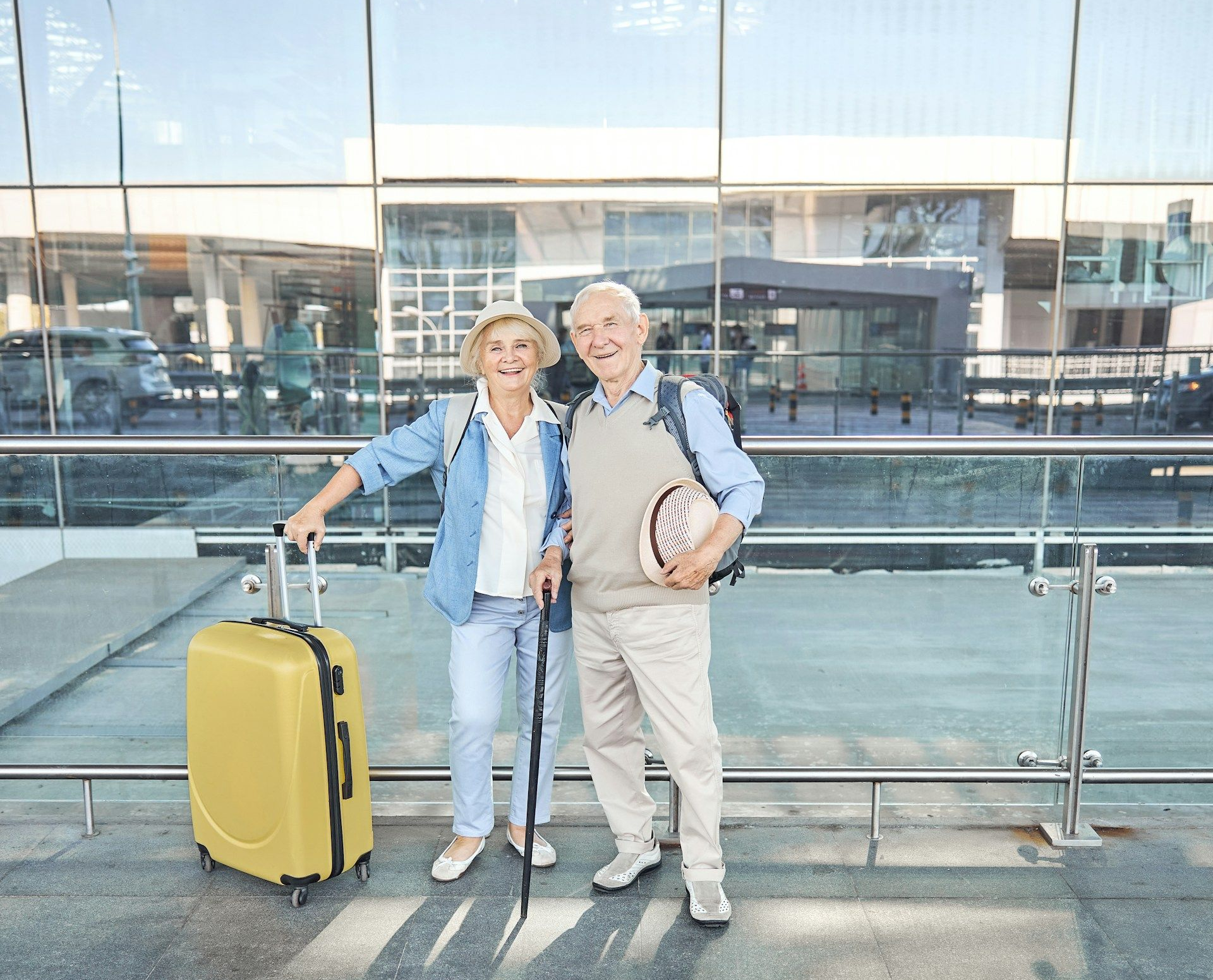 Elderly couple smiling, posing near airport entrance. Woman with yellow suitcase, man with cane.