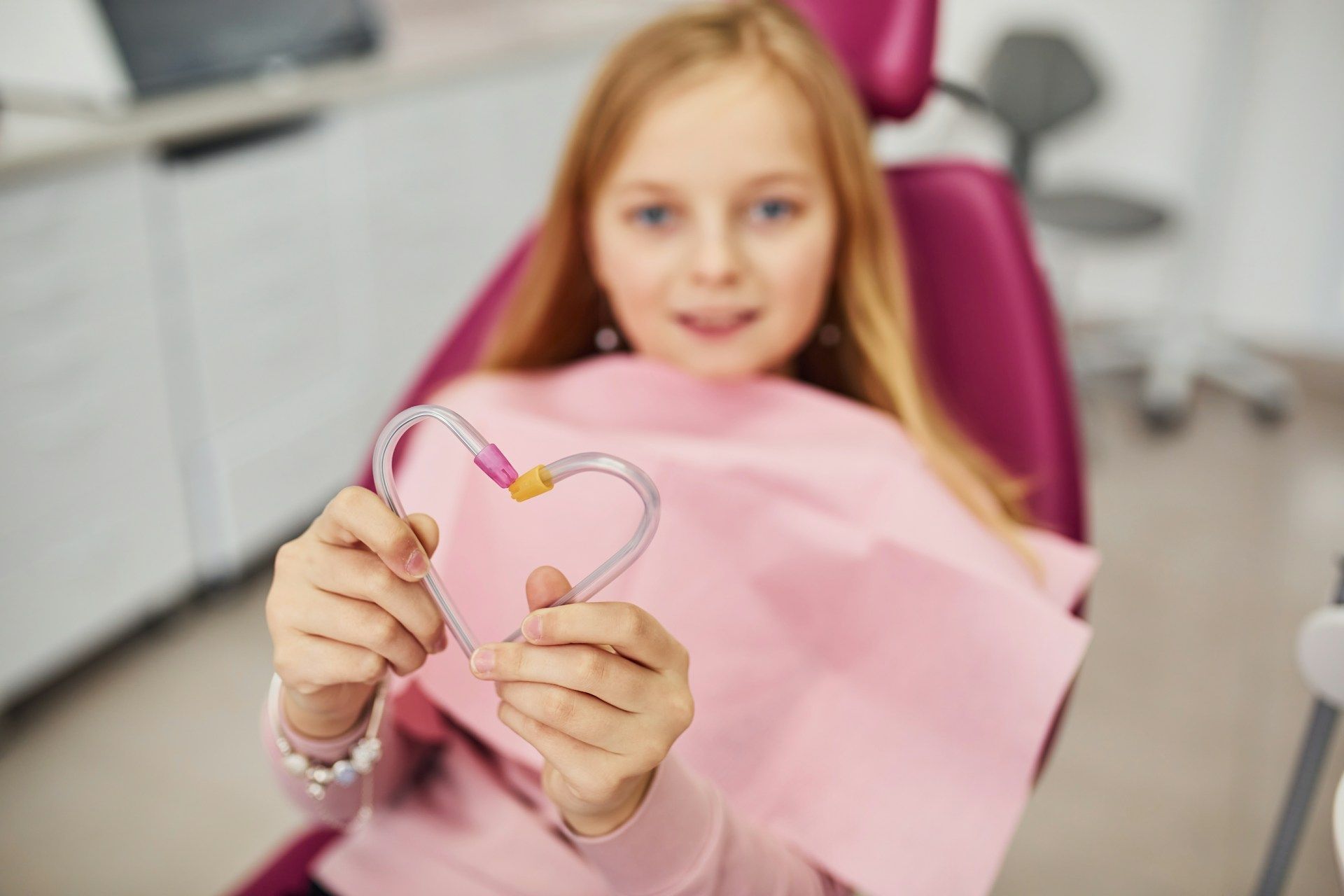 Girl in dental chair making a heart with dental suction tubes. Pink bib and chair.