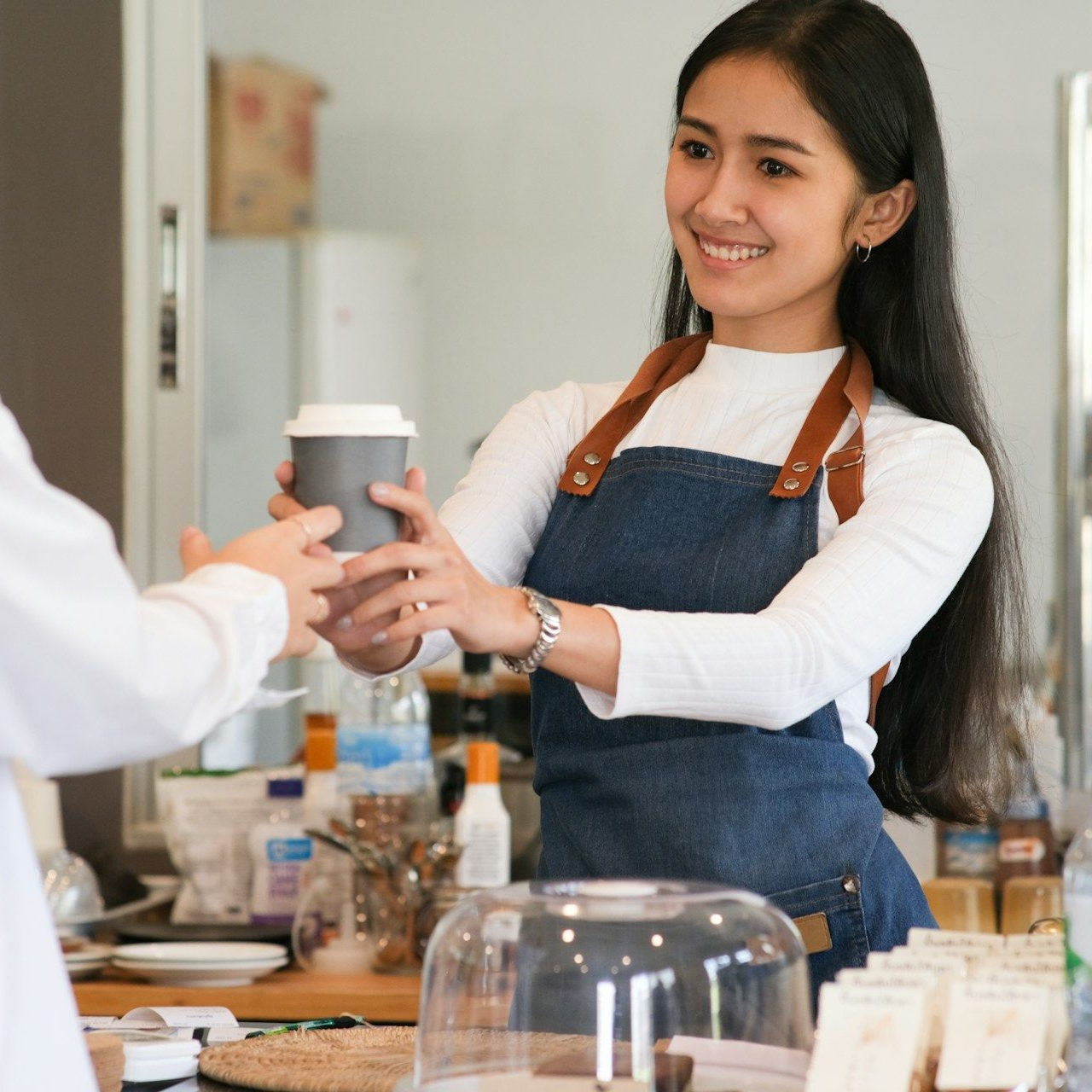 Barista handing a coffee cup to a customer; cafe setting, smiling.