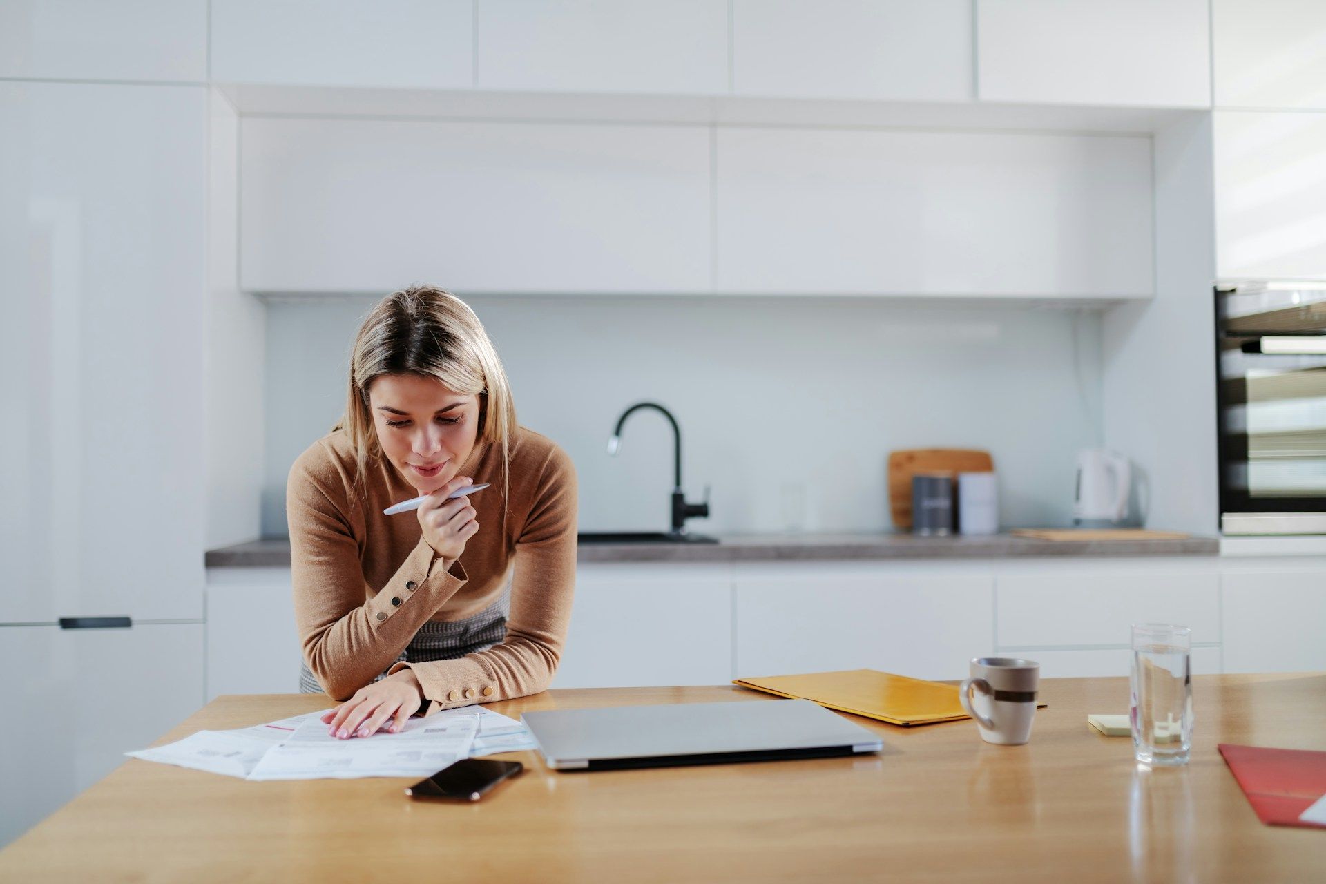 Woman in kitchen, studying documents, with laptop and coffee cup on table.