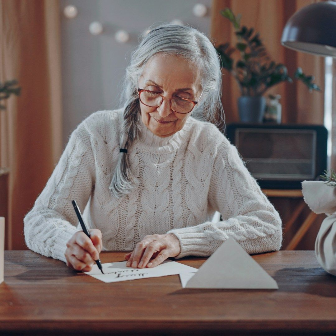 Elderly woman with glasses writes at a wooden desk, inside a room with plants.