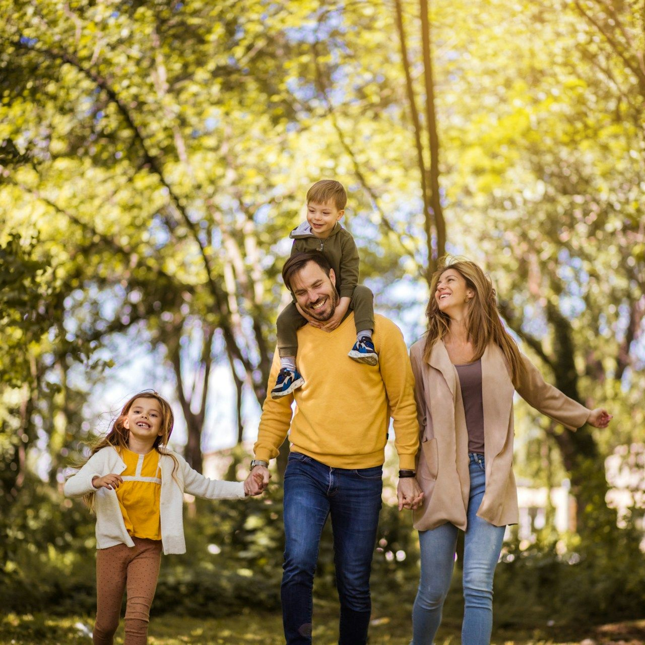 Family of four smiling, walking together in a park. Father with son on shoulders, mother and daughter holding hands.