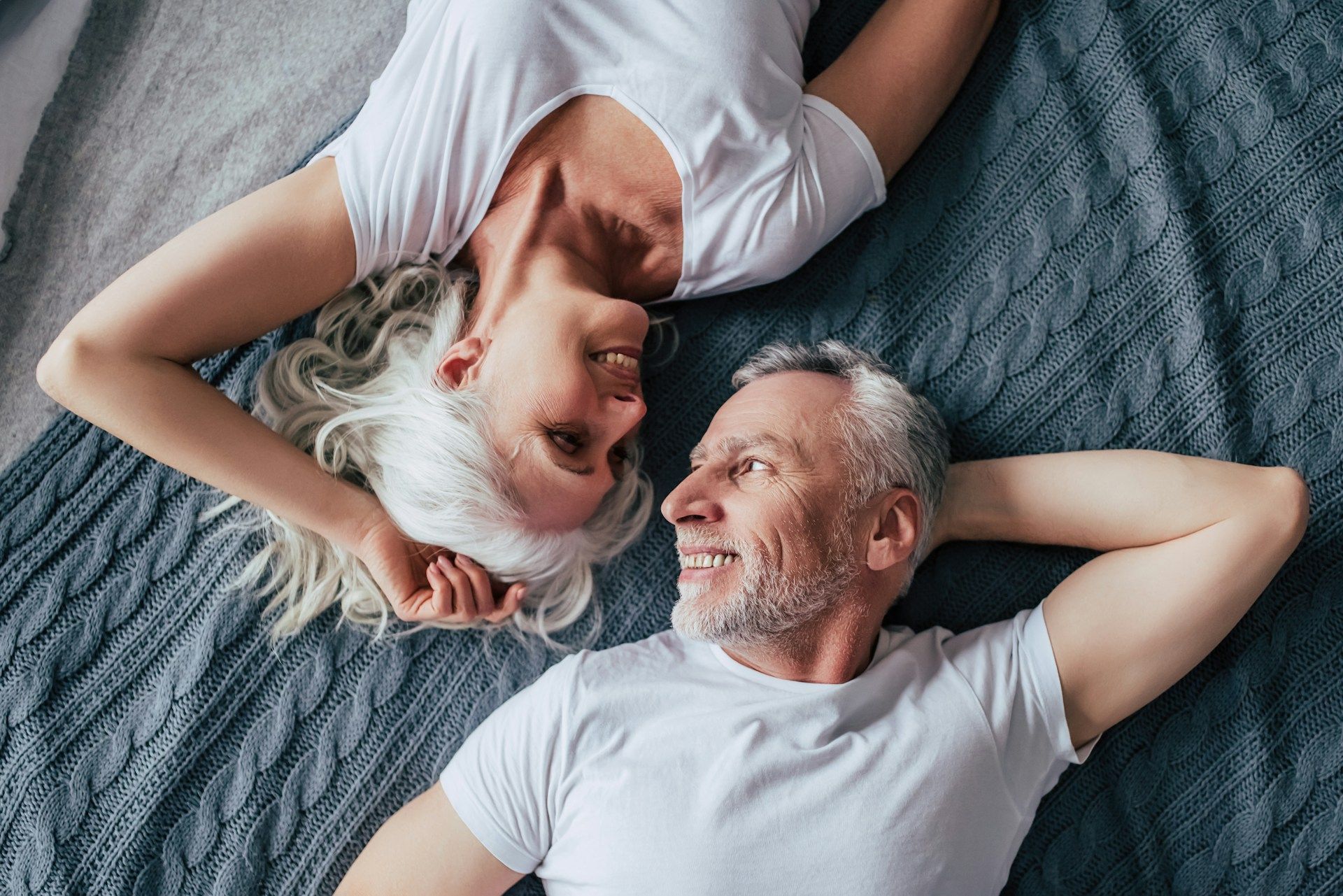 Couple, gray-haired, lying on bed, smiling at each other, wearing white shirts, top-down view.