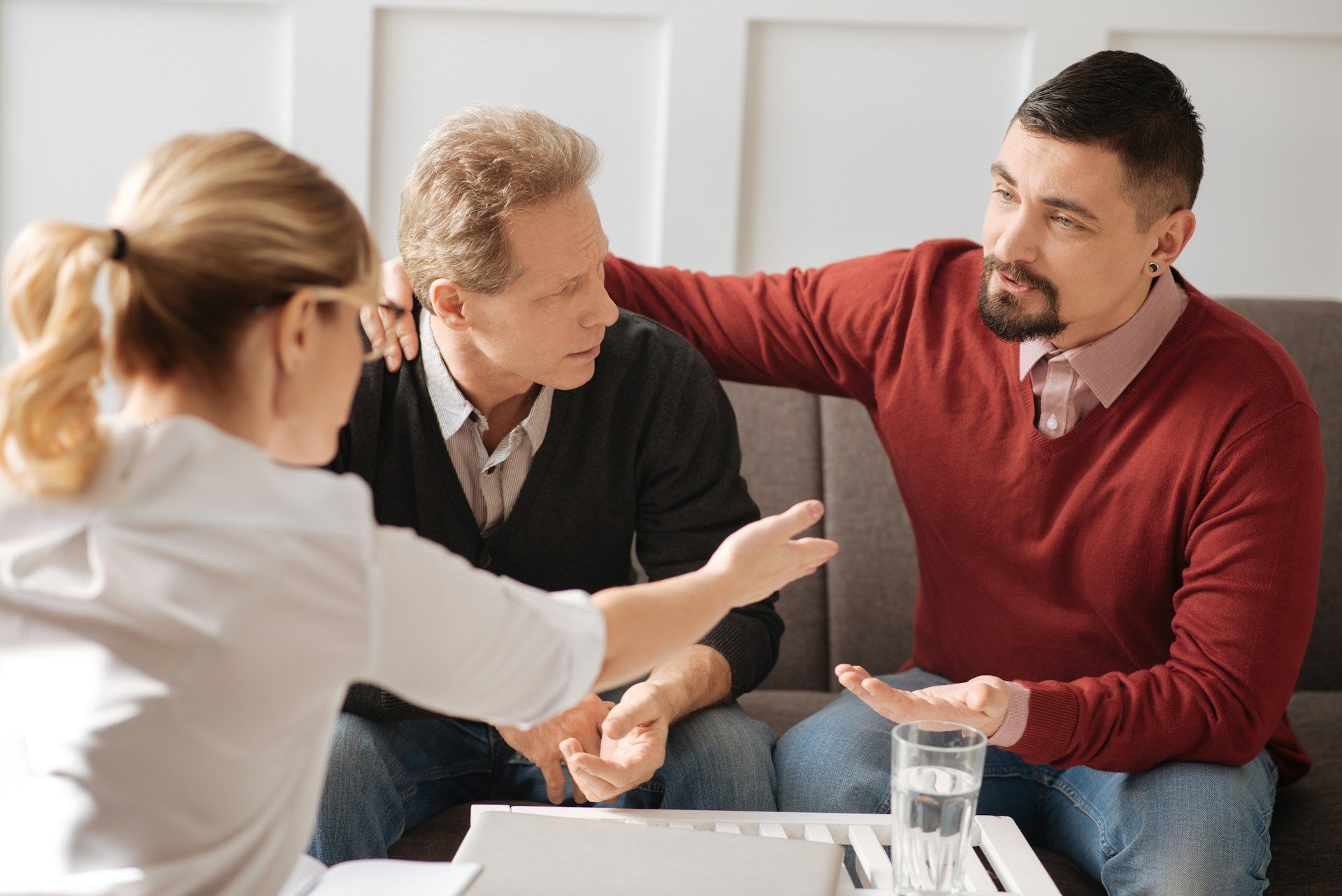 A therapist with a couple: Man comforting another man. They are sitting on a couch.