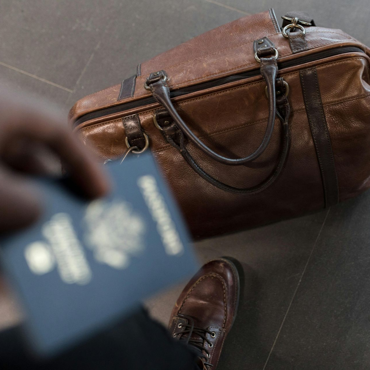 Person holding a passport near a brown leather travel bag and brown leather shoe, ready to travel.