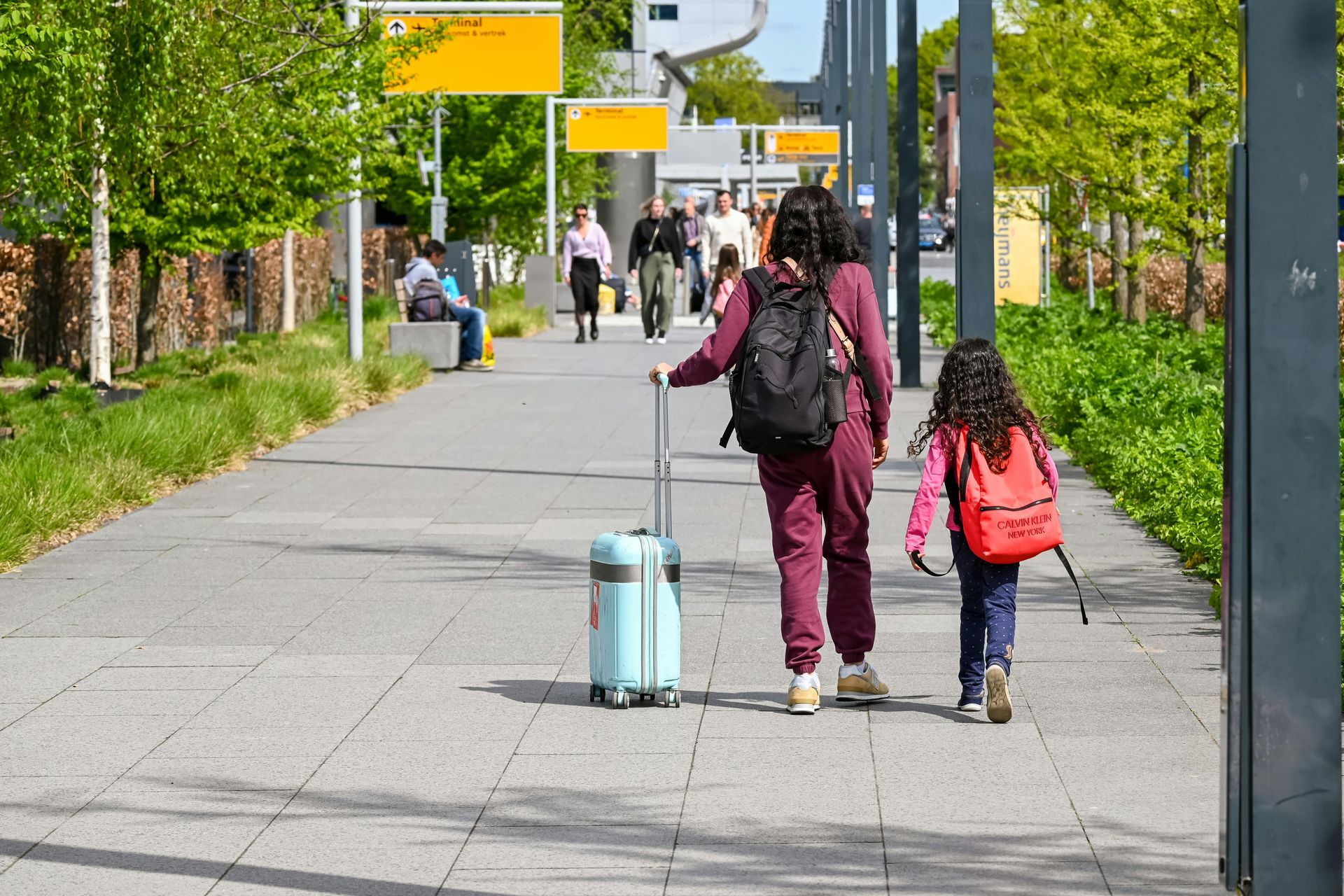 Woman and child walk with luggage on a paved path; trees and people in the background.