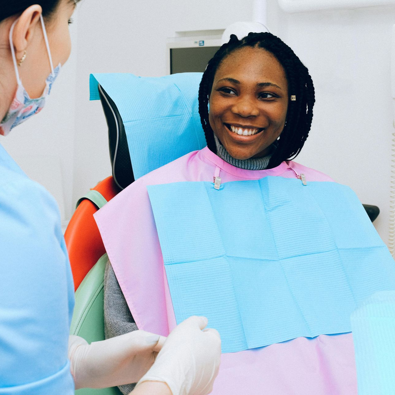 A dentist and a smiling Black woman in a dental chair; dentist in blue, patient in pink bib.