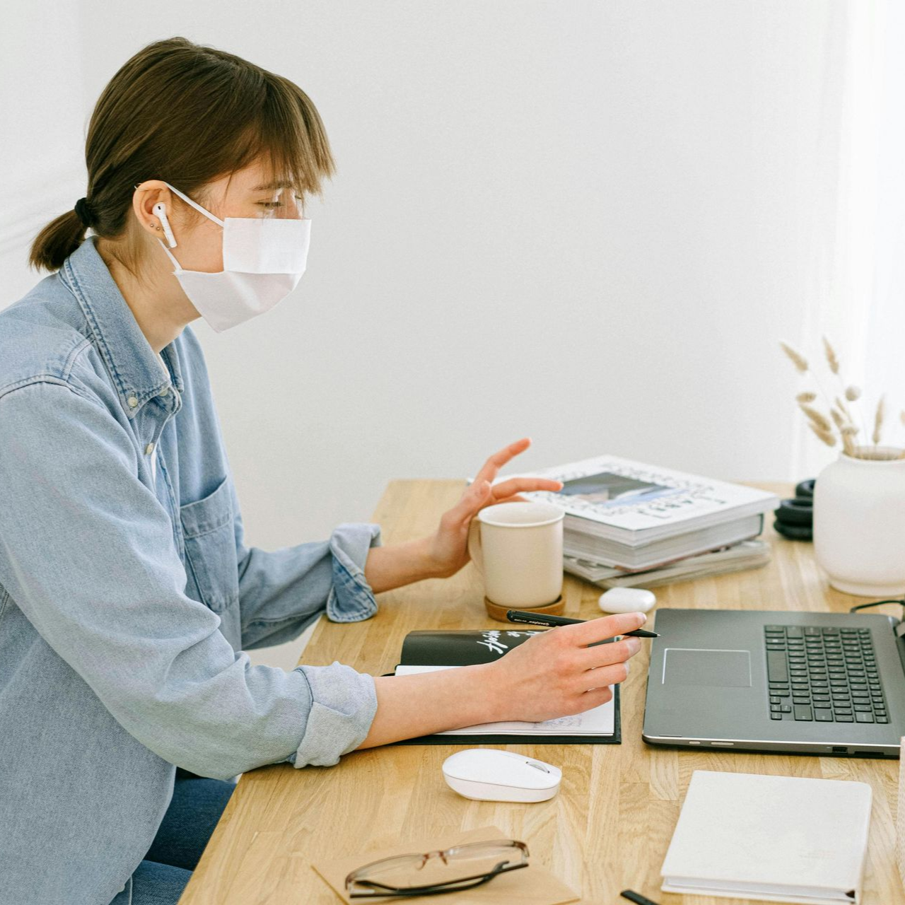 Woman in mask working on laptop at a desk, holding a mug, with a notebook and books.