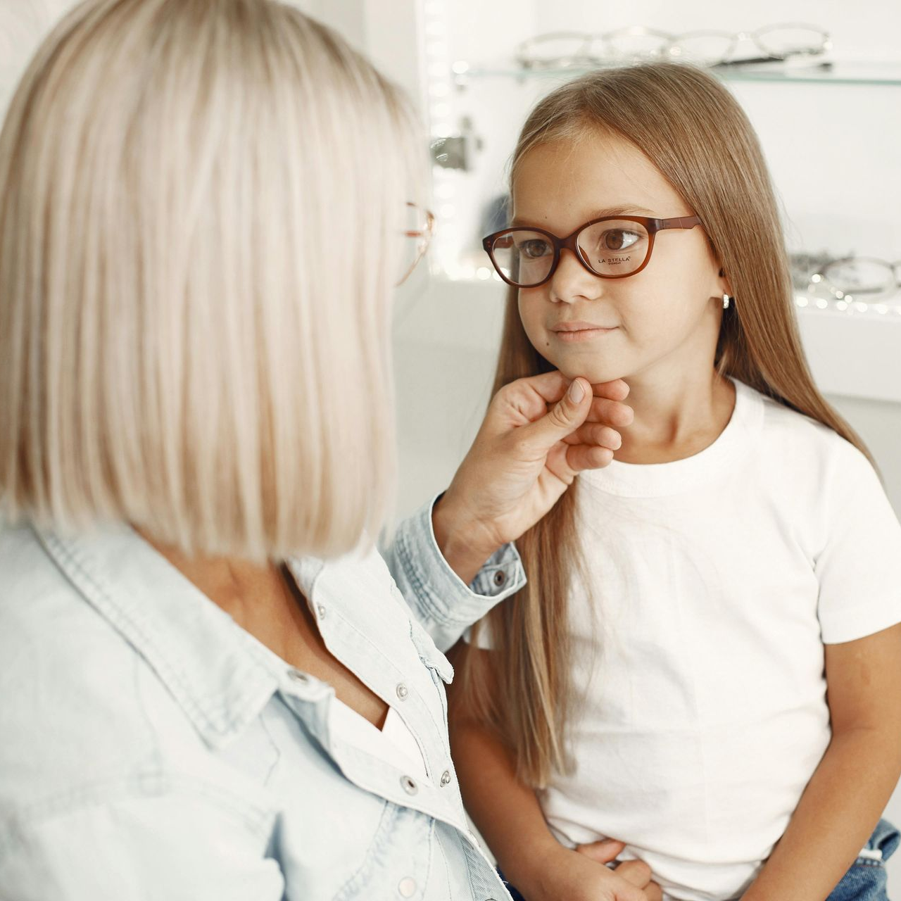 Woman adjusts eyeglasses on a young girl in an eye doctor's office. Both are fair-skinned.