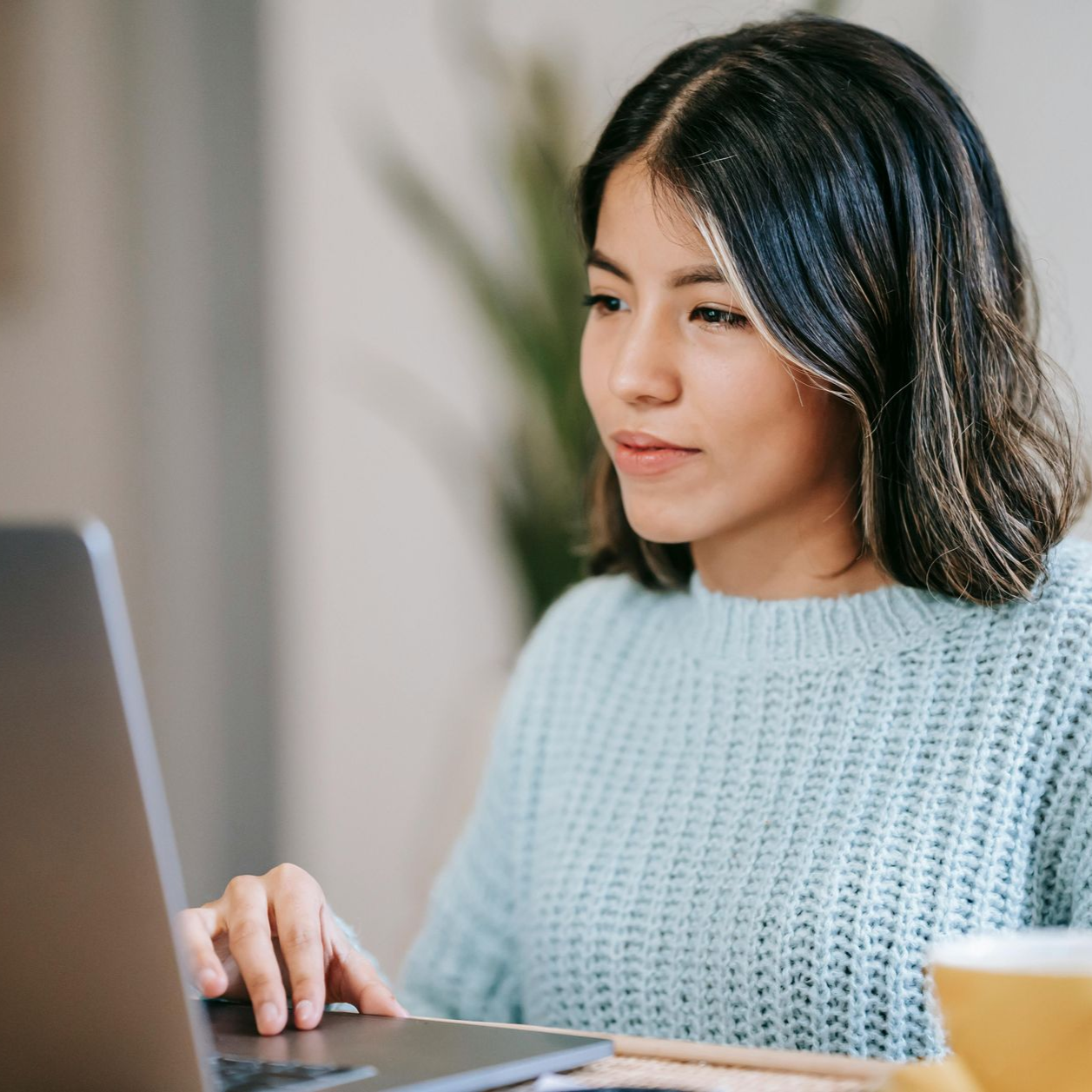 Woman with dark hair using a laptop, indoors, wearing a light blue sweater, looking focused.