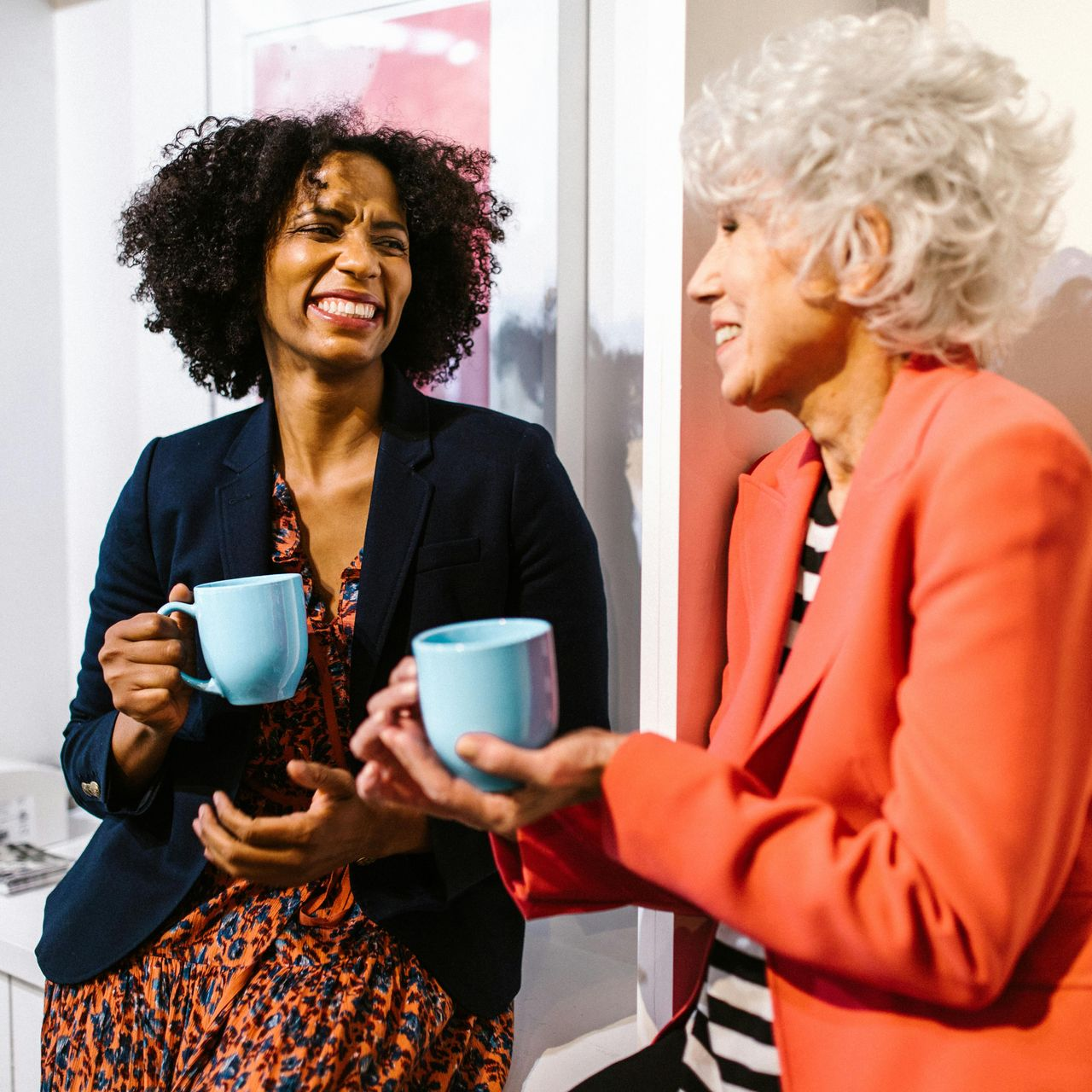 Two women in office setting, laughing, holding blue mugs. One in navy blazer, one in red.
