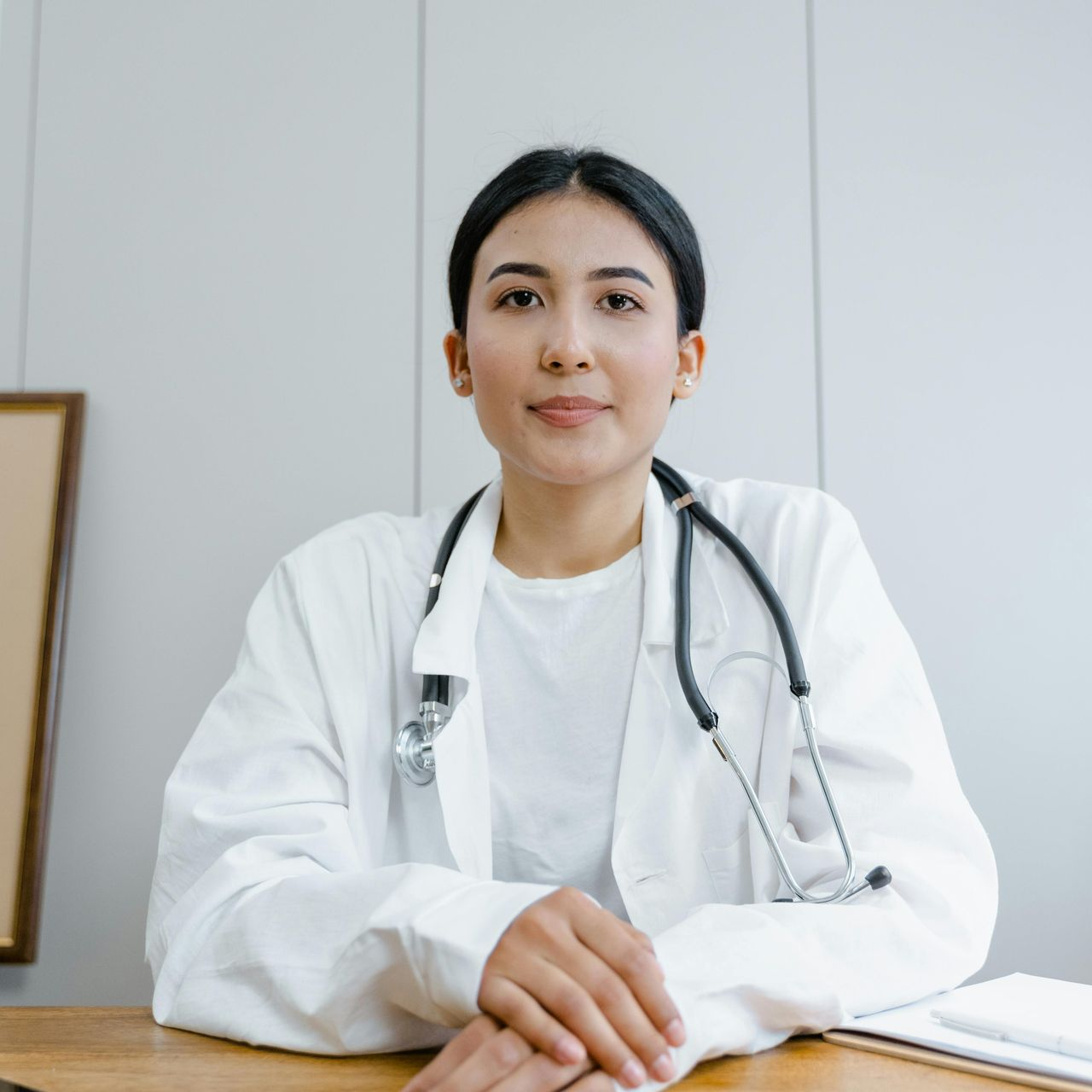 Woman doctor in white coat with stethoscope, seated at desk, smiling, indoors.