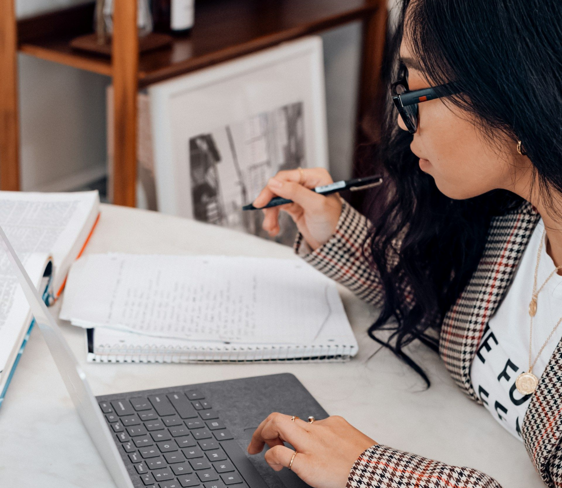 Woman with glasses working on a laptop, taking notes with pen, in a home office.