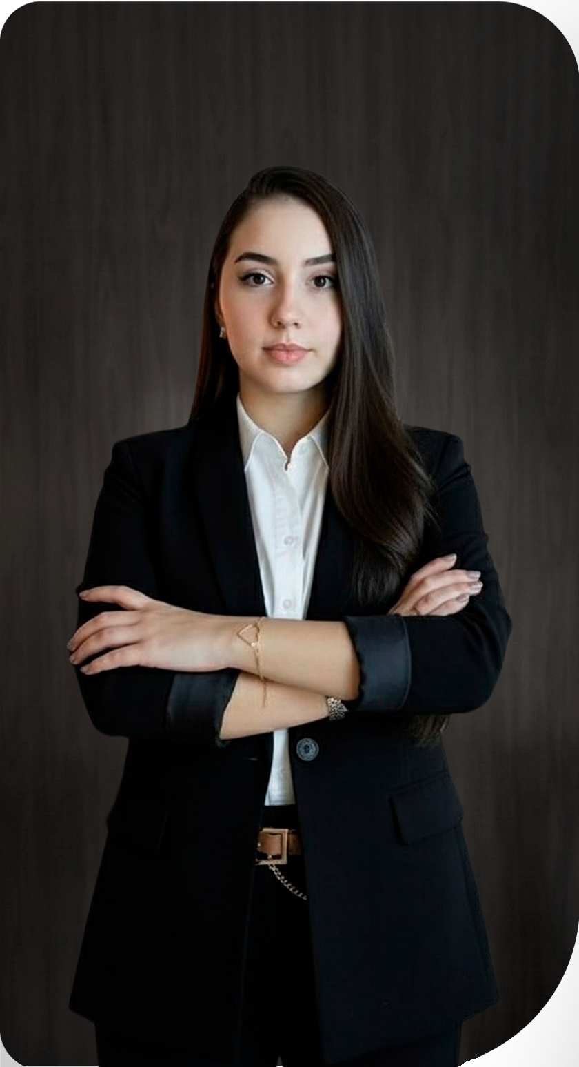 Mulher de blazer preto com braços cruzados, camisa branca, cabelo castanho, fundo escuro.