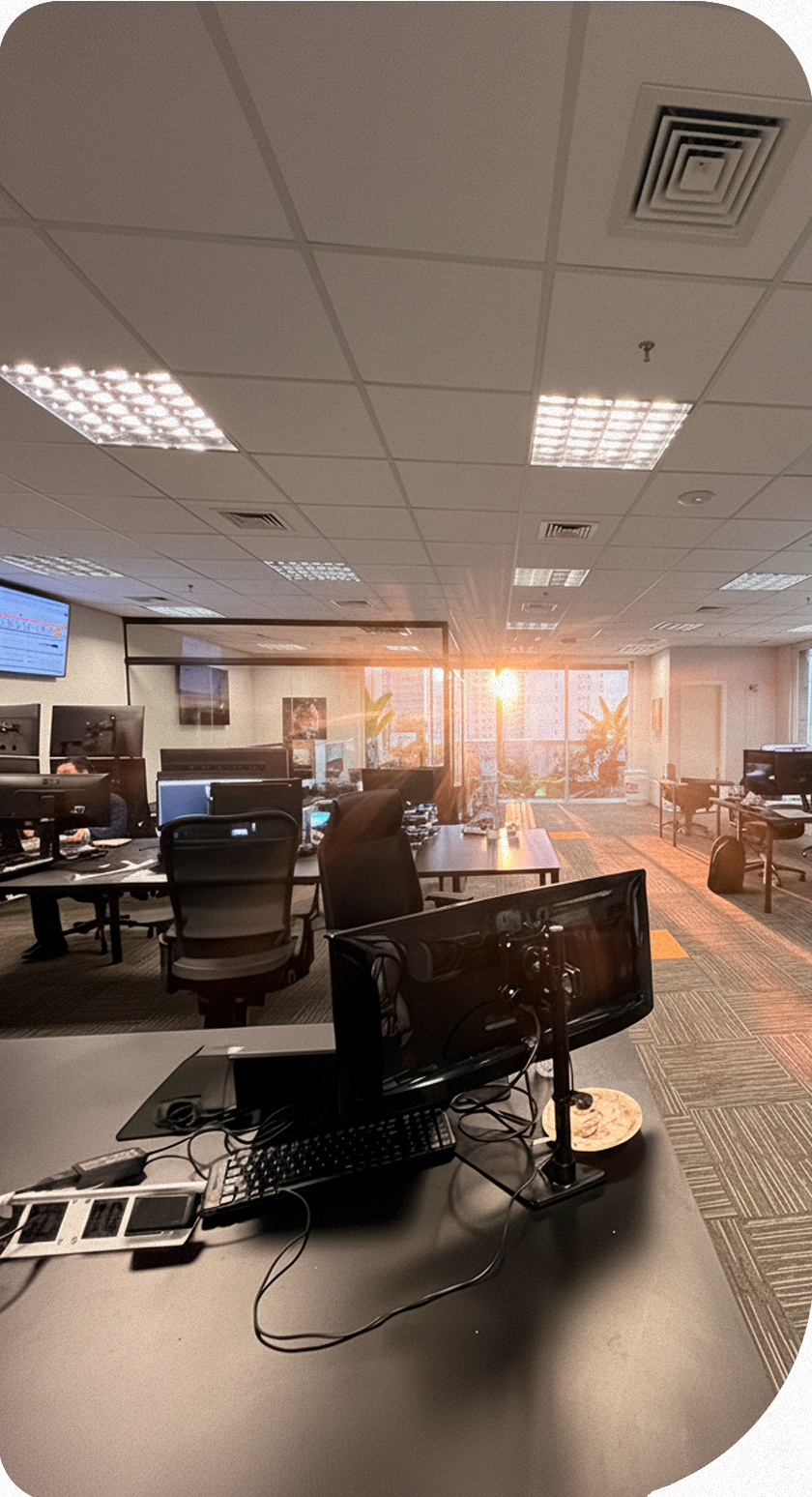 Espaço de trabalho interno com mesas, computadores e cadeiras. A luz do sol entra pelas janelas.