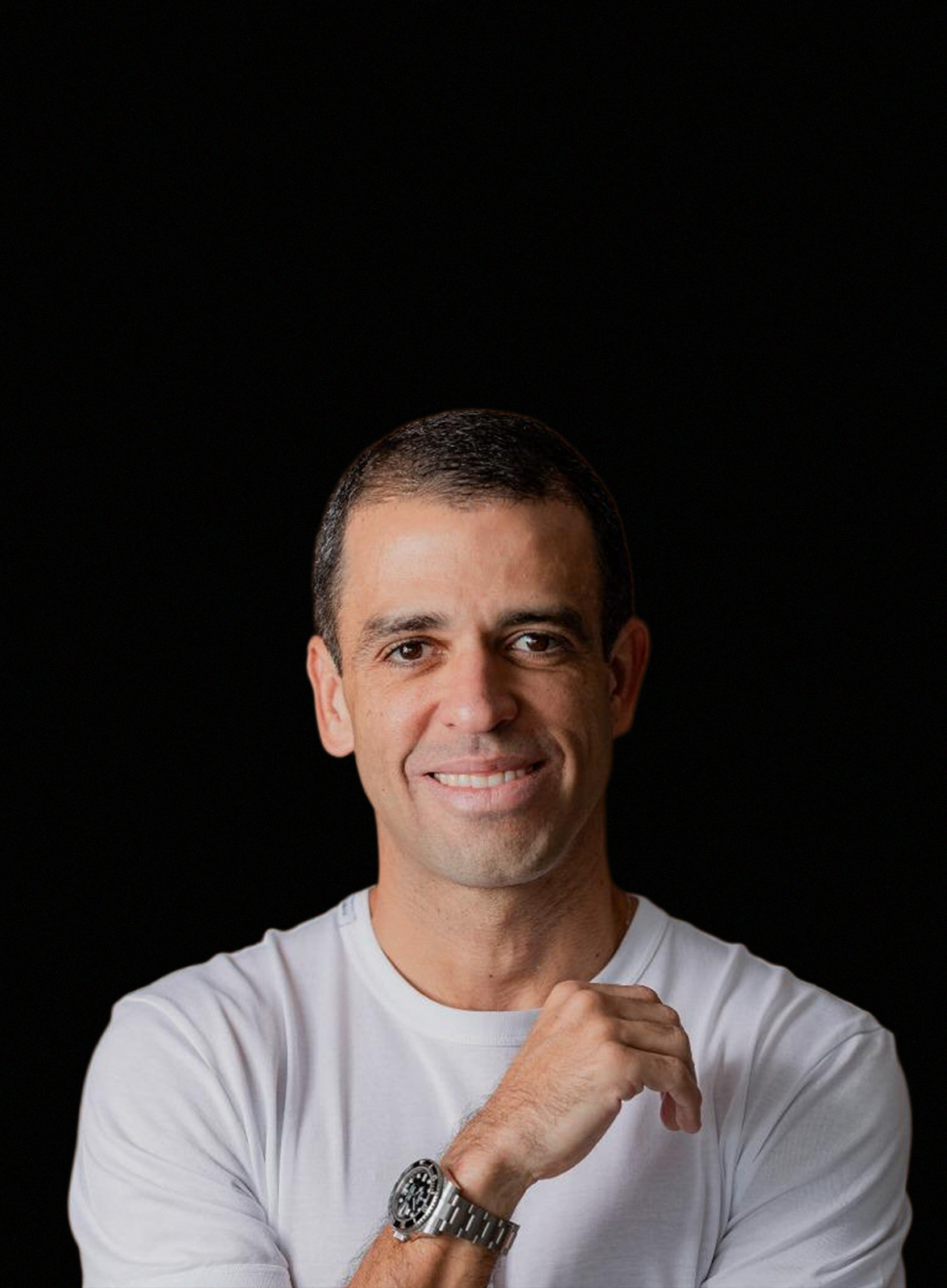 Homem de camisa branca sorrindo, com a mão no rosto, contra um fundo preto.