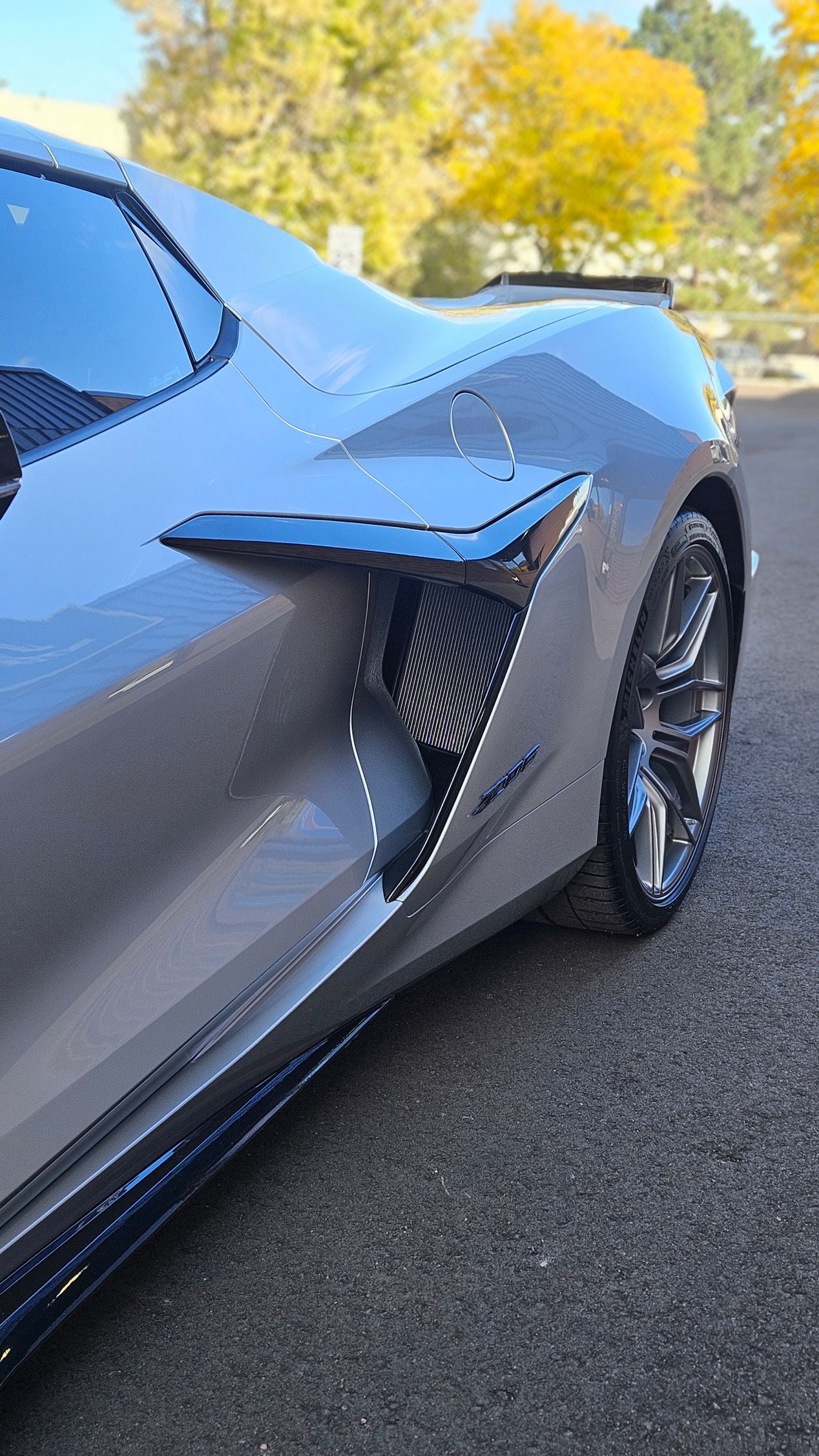 Silver sports car with black vents parked on asphalt; fall foliage in background.