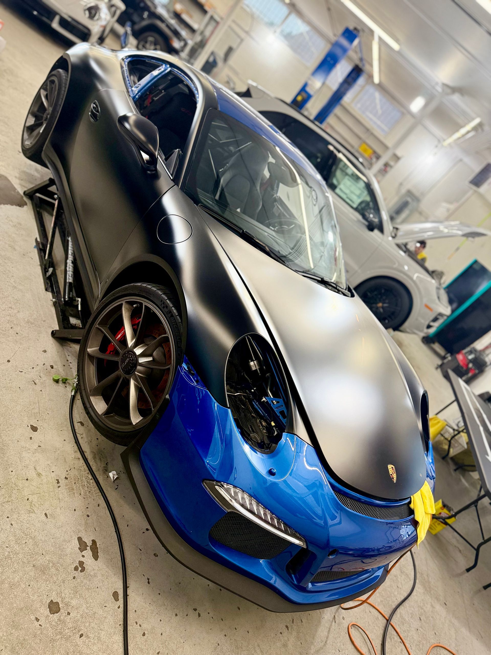 Blue and black Porsche being worked on in a garage; front end in blue, matte black body.