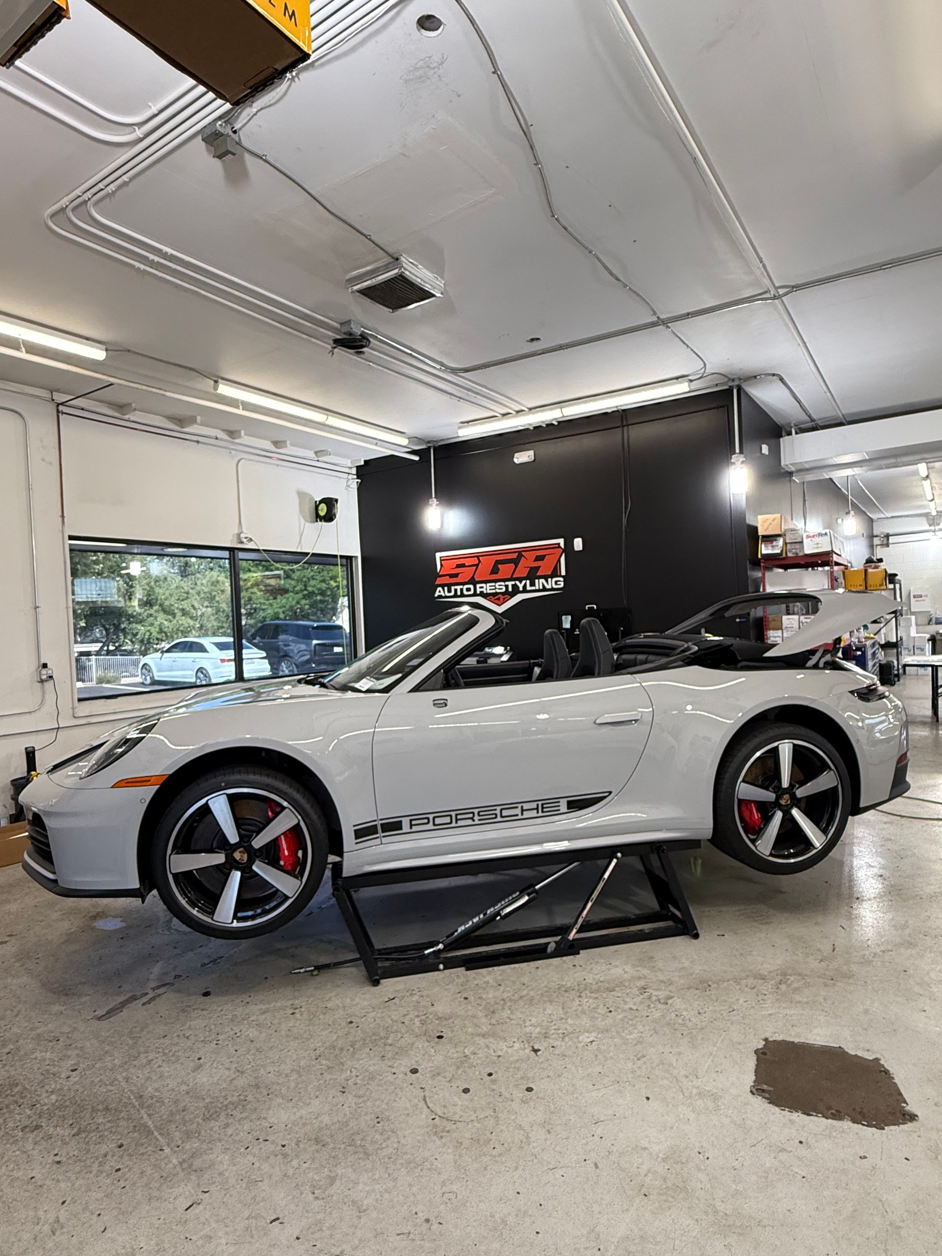 A light gray Porsche convertible on a lift inside a workshop. Red brake calipers, black wheels, and a black backdrop.