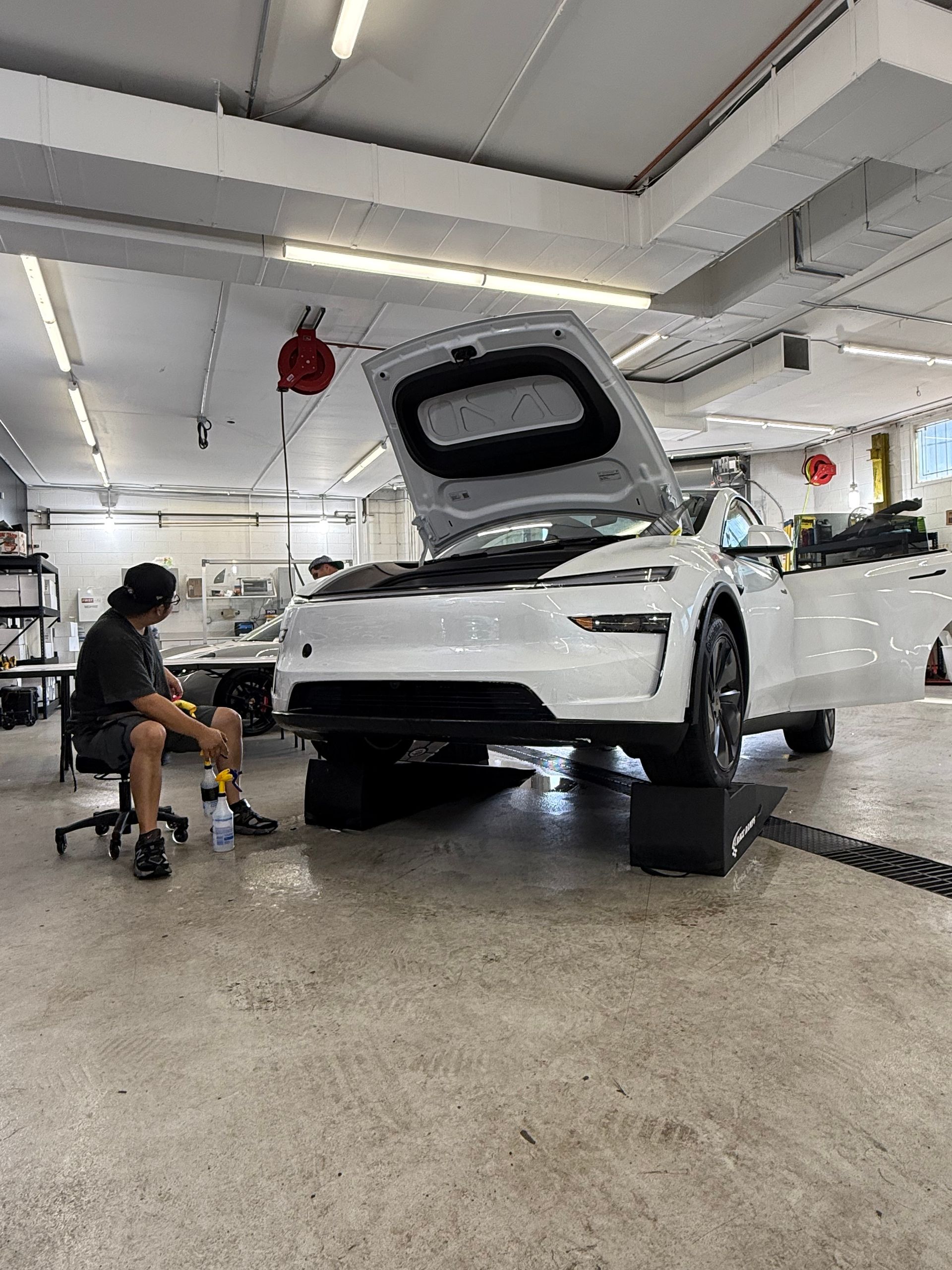 A white car with the hood open, being worked on in a garage. A person sits nearby.
