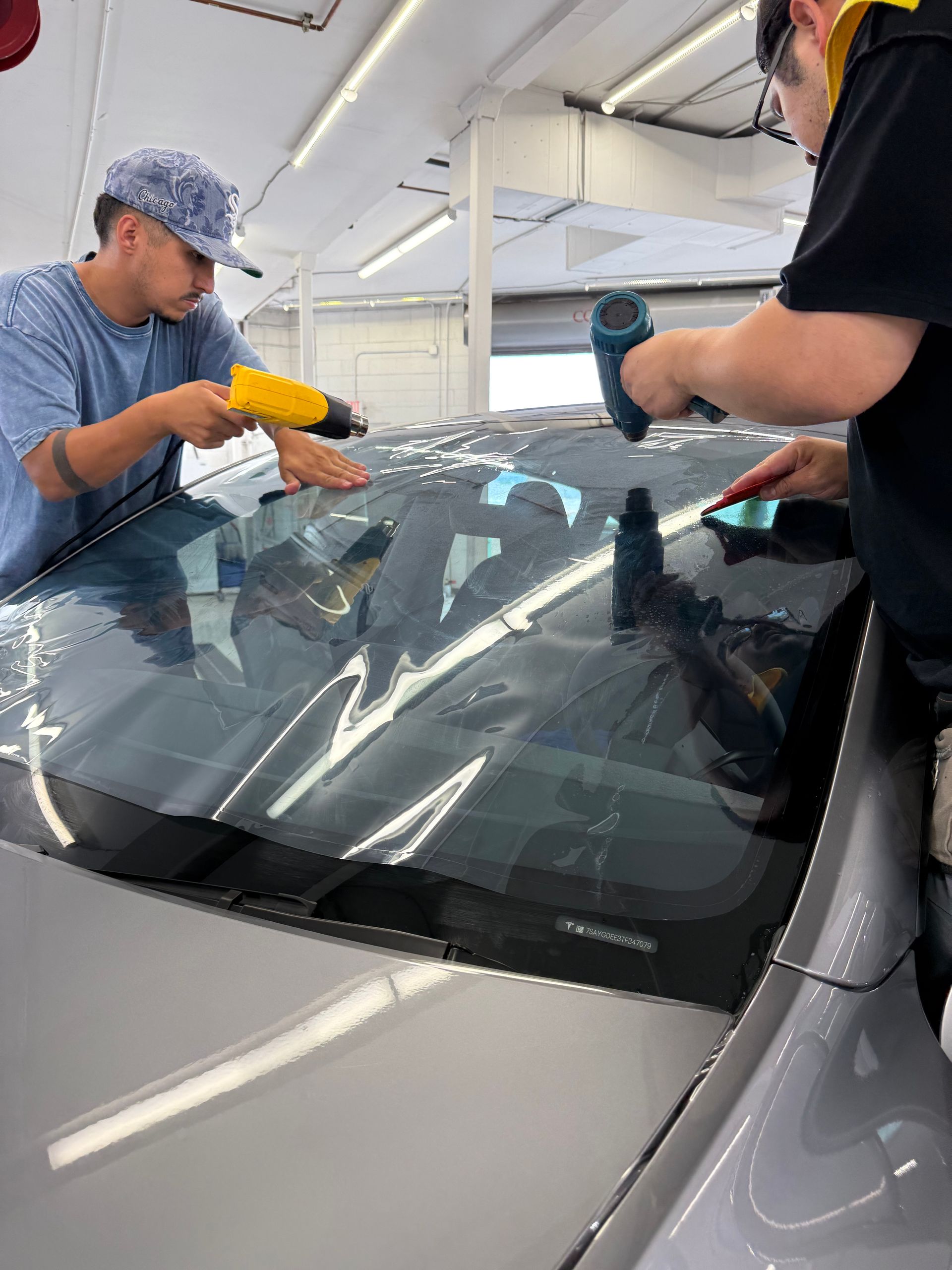Two people applying tint to a car's rear window with heat guns in a shop.