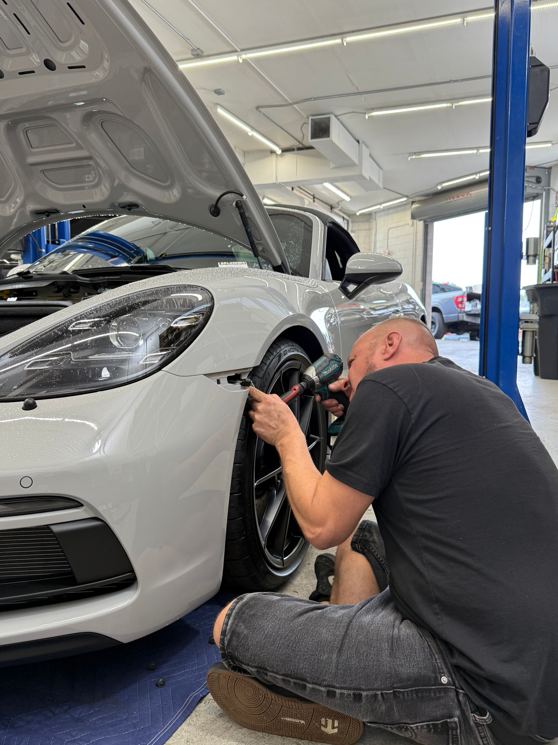 Mechanic working on the front of a silver car with the hood up inside a garage.