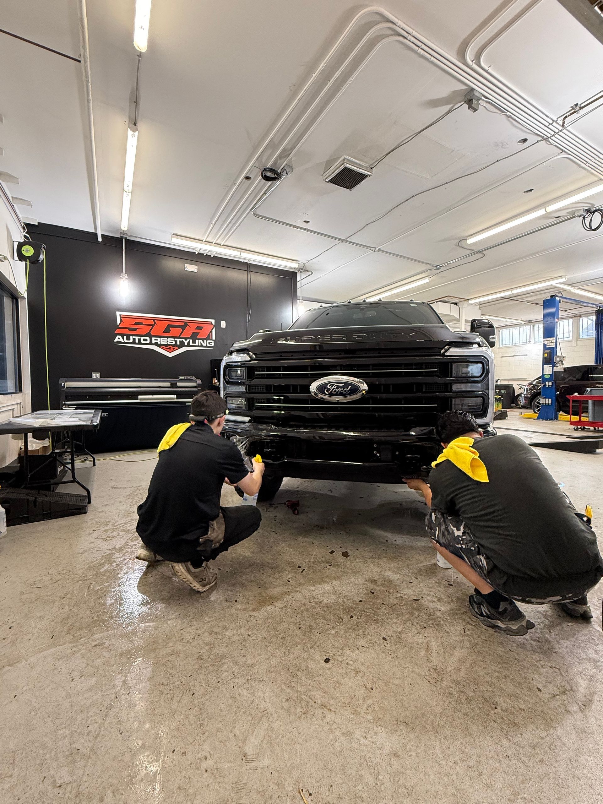 Two workers installing a black front bumper on a black Ford truck inside a garage.