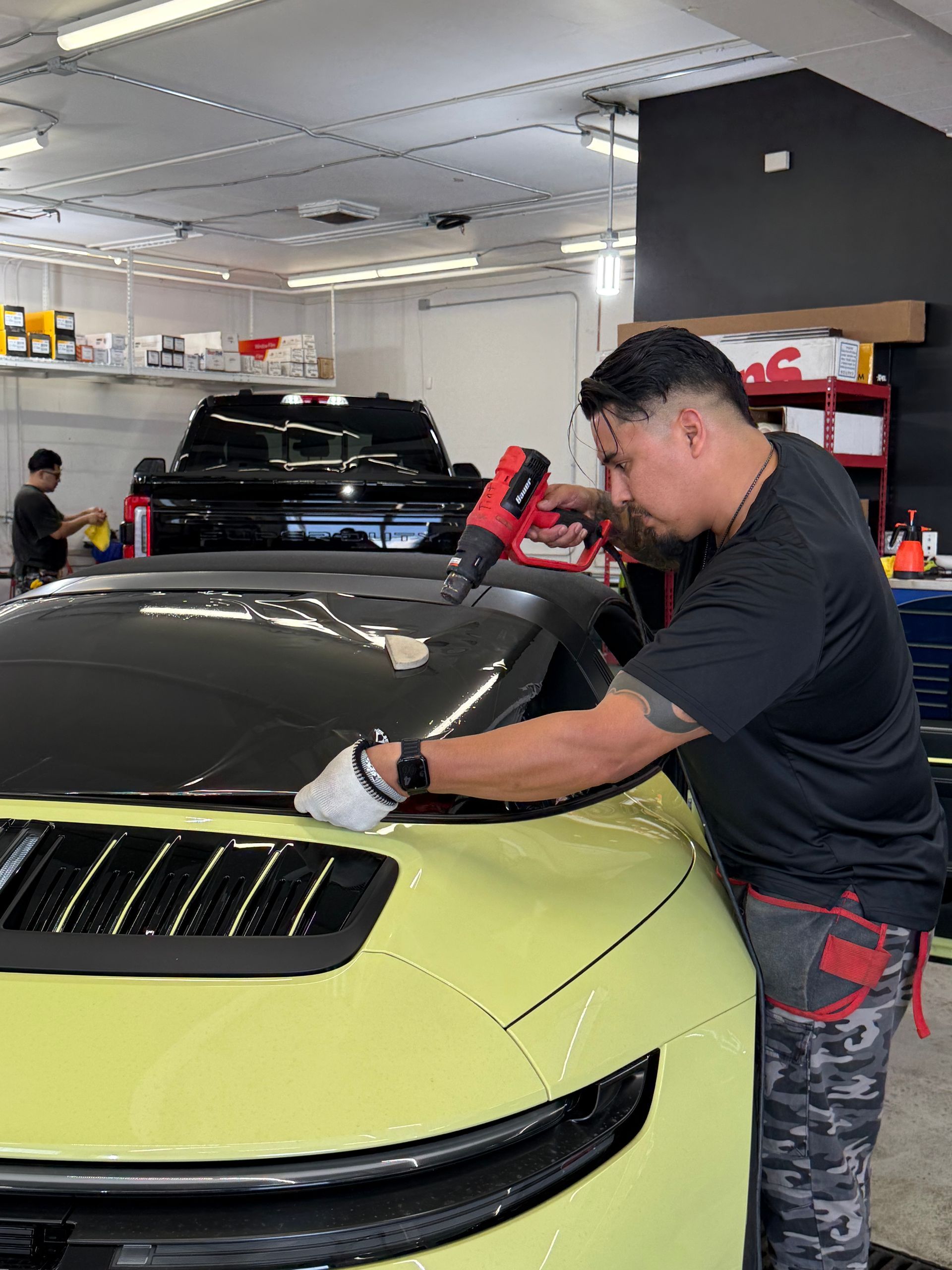 Man applying film to a yellow car's hood in a garage, using a heat gun. Black truck and shelves visible.
