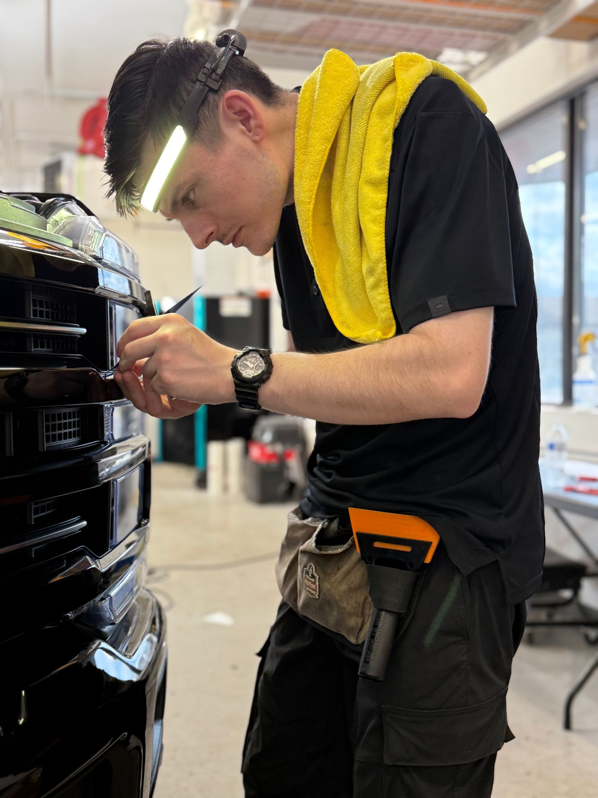Person working on a car grille, wearing a headlamp and carrying tools. Yellow towel around neck. Indoor setting.