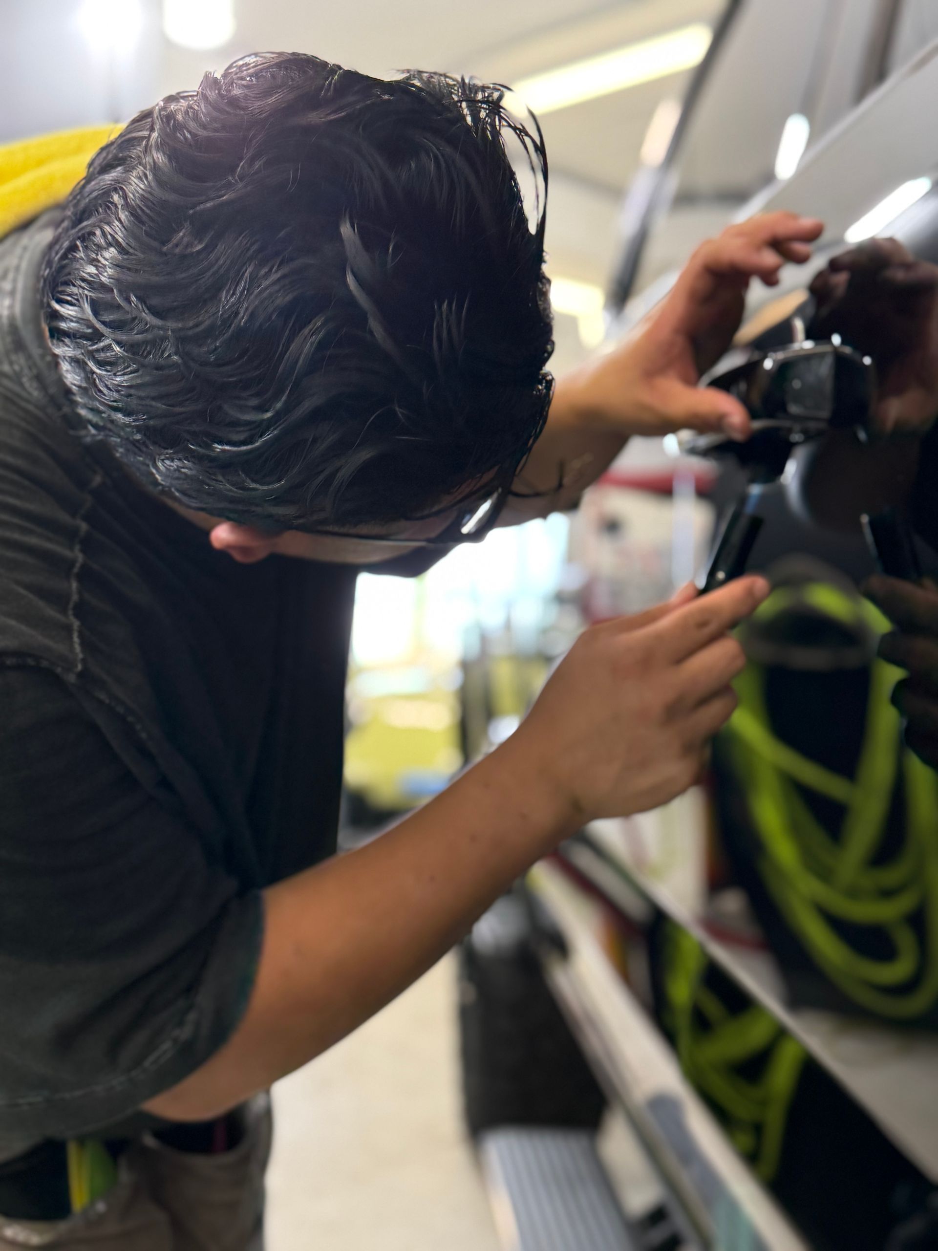 Person working on a car door, likely applying a film. Black car, gray shirt, bright interior.