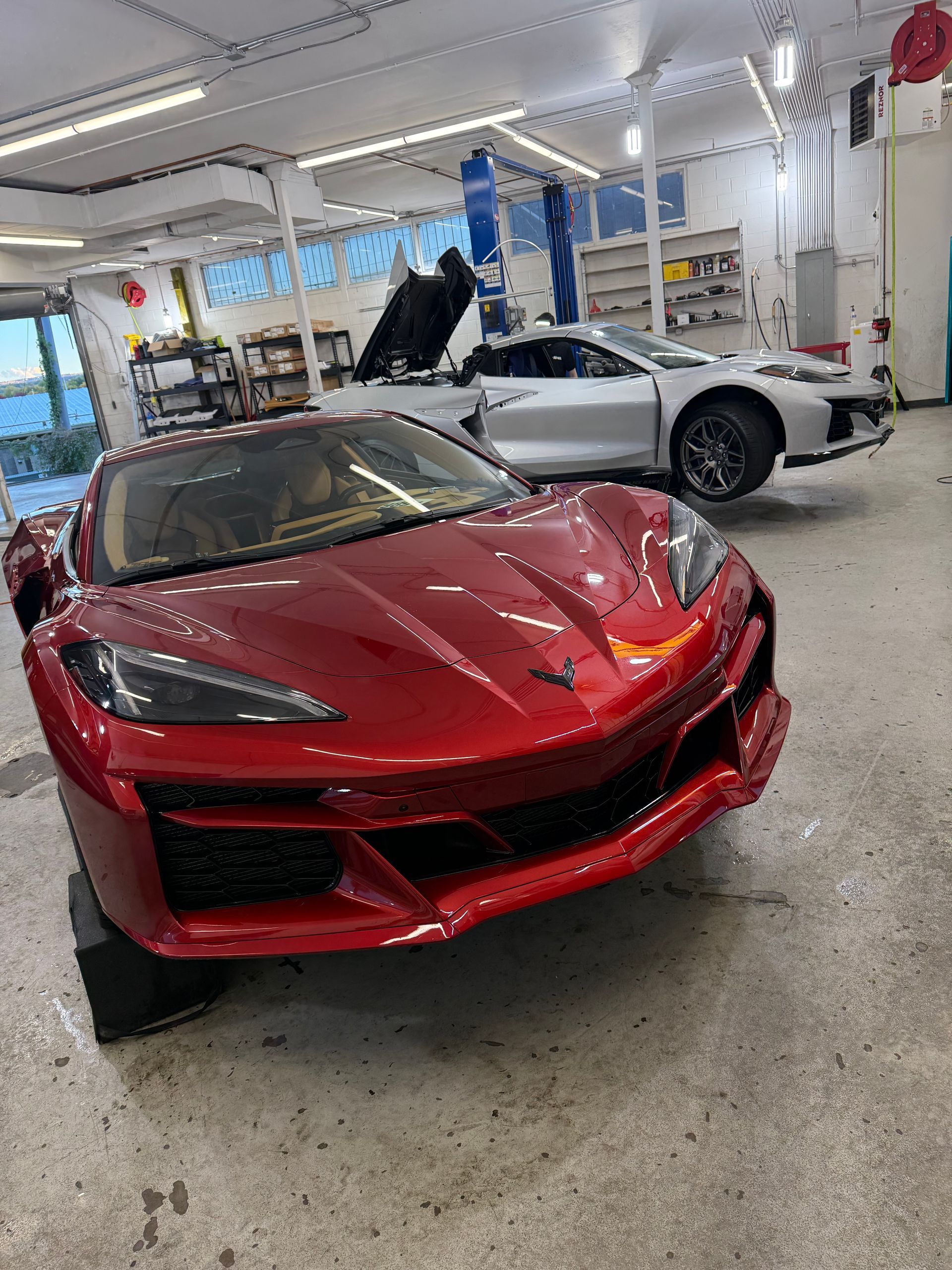 Red Corvette sports car in a garage, another silver Corvette in the background, doors open.