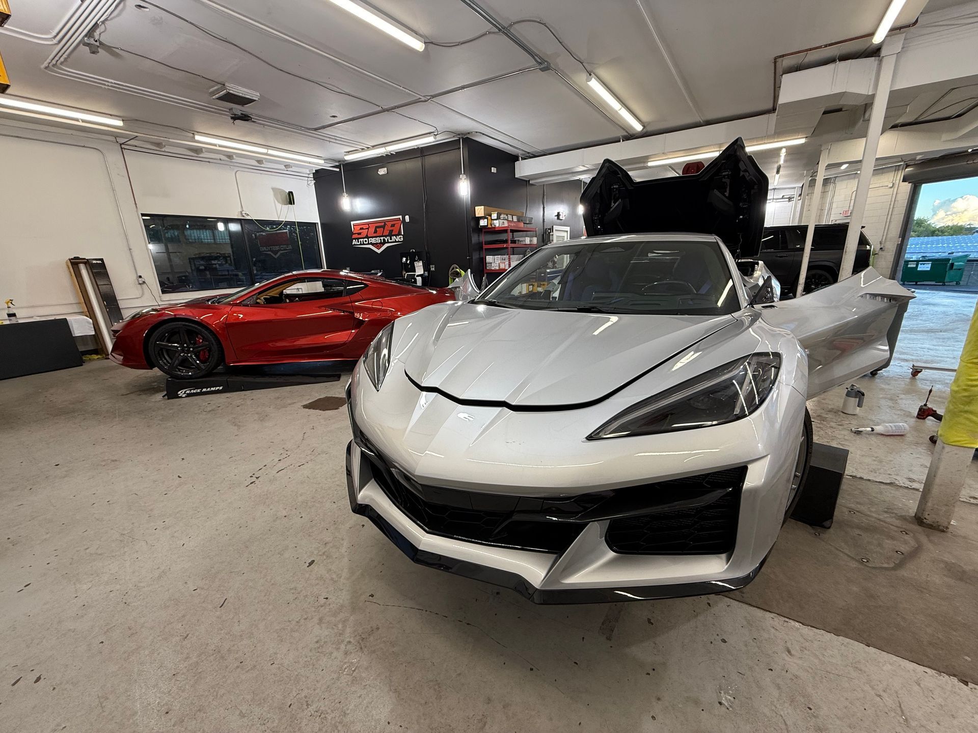 Silver sports car with open doors in a garage, red sports car in the background.