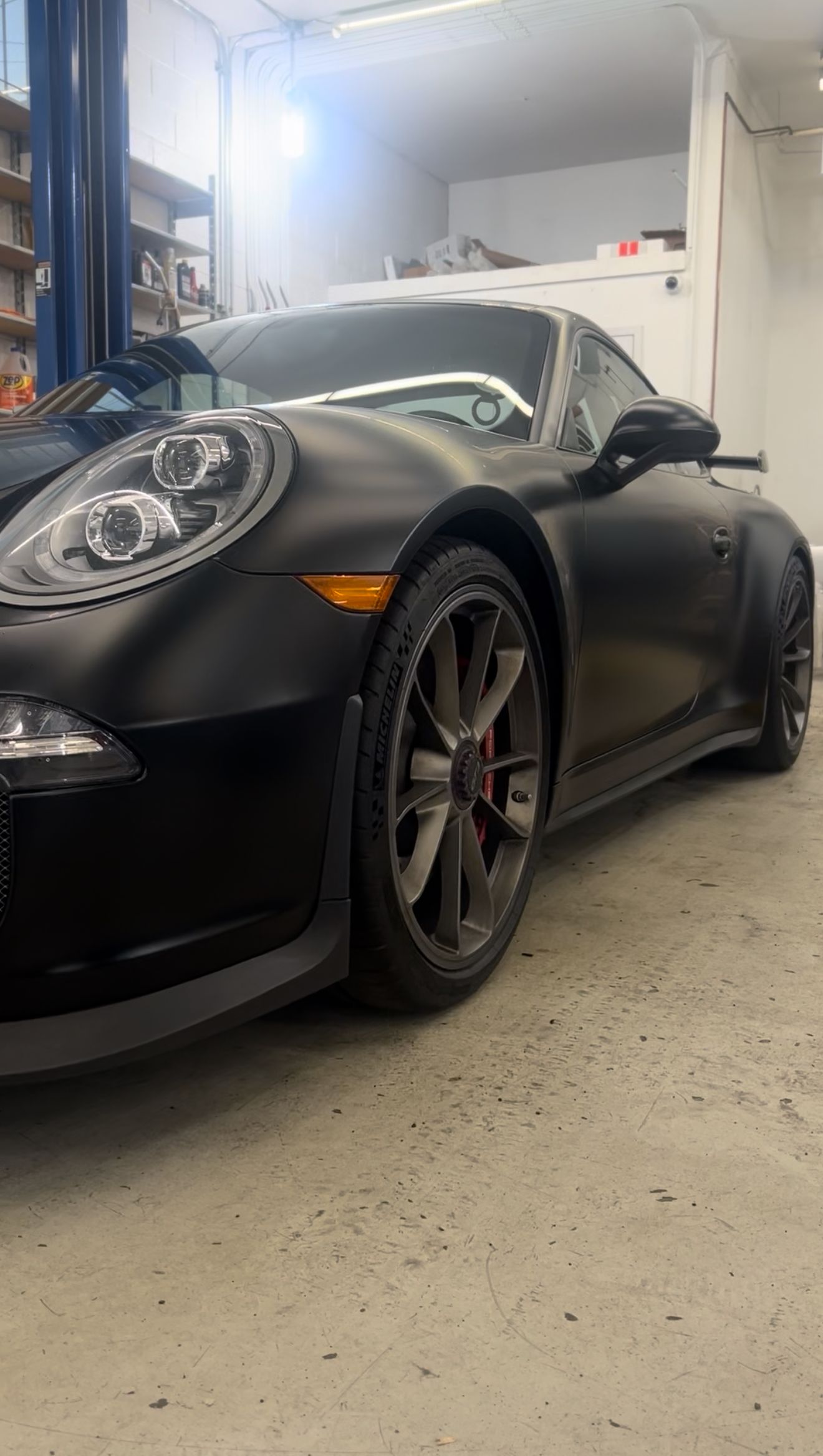 Black Porsche sports car in a garage, parked at an angle. Matte finish, gray wheels, orange side marker.
