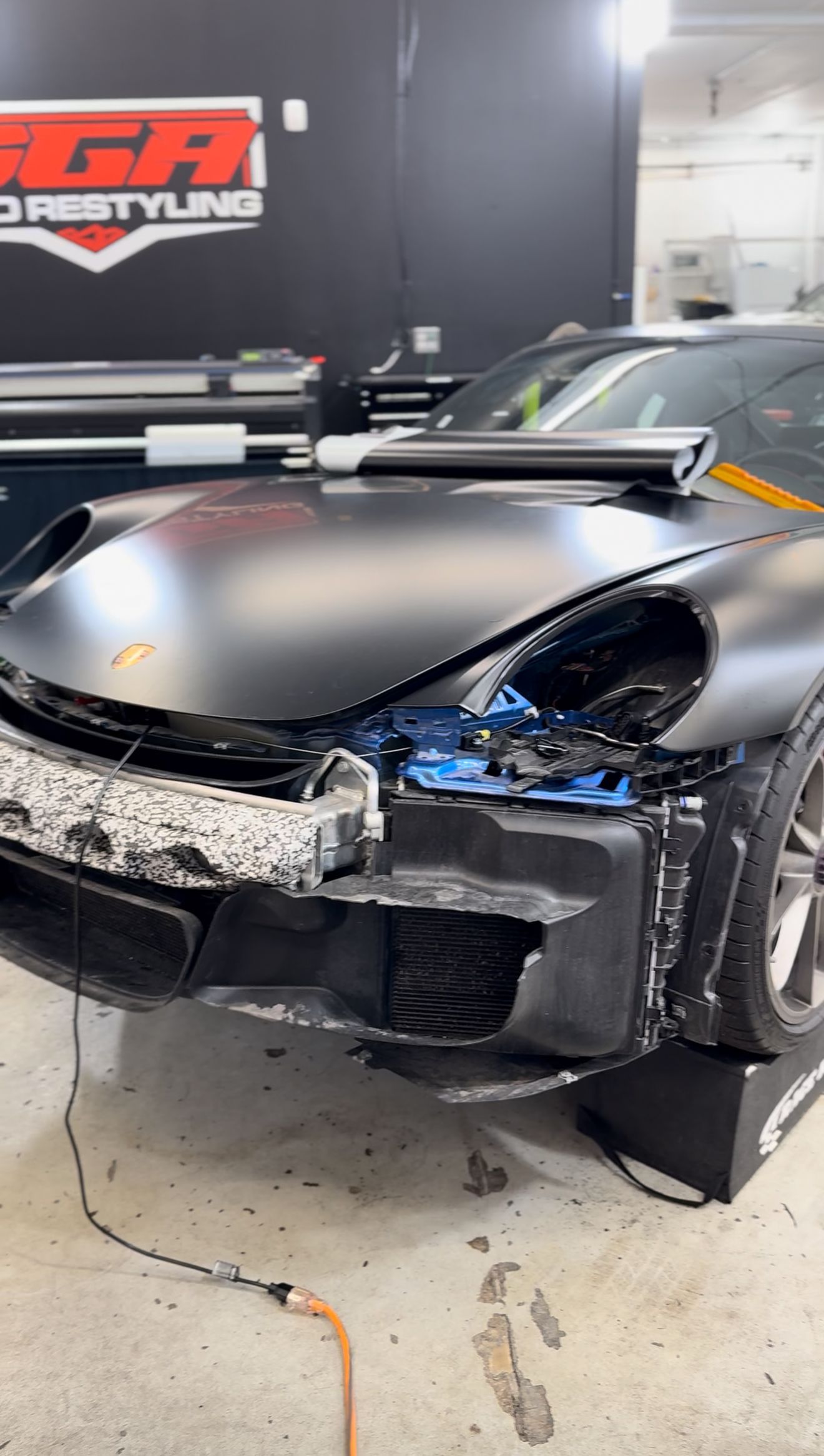A black sports car partially wrapped in matte vinyl, with its front bumper disassembled in a shop.