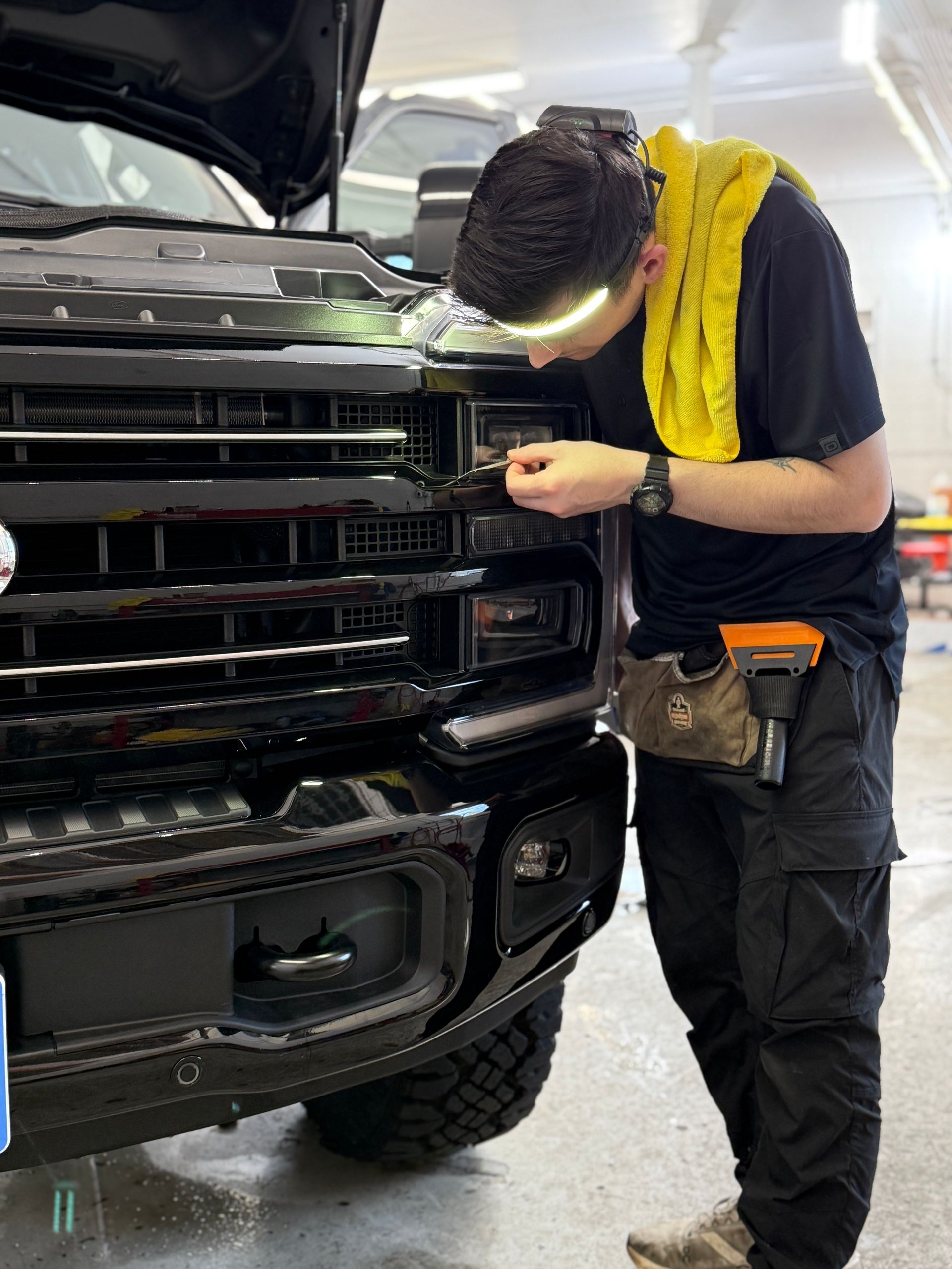 Man working on truck's front grill with a headlamp, outdoors, black truck, black grill.