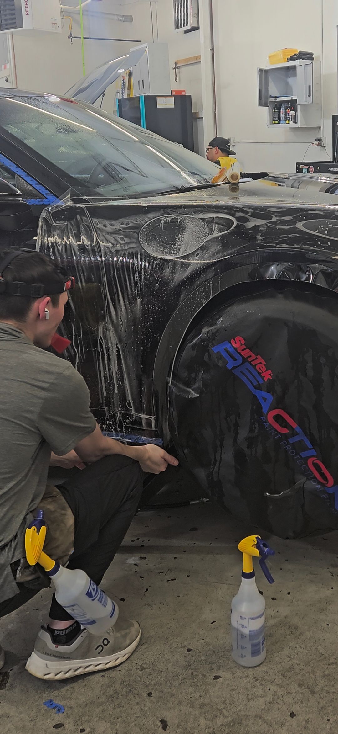 A person applying a protective film to a black car, in a workshop setting.