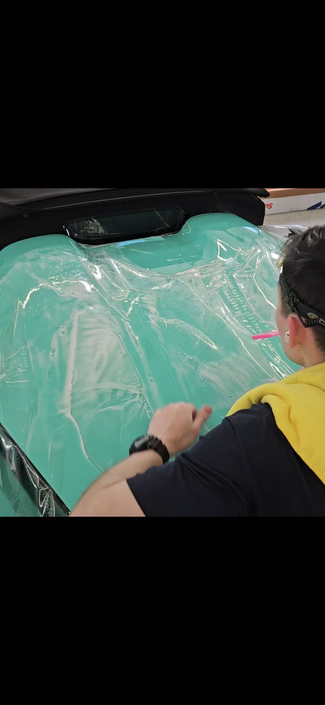 Person working with a turquoise substance, possibly applying a clear film. The person wears a yellow top and a black watch.