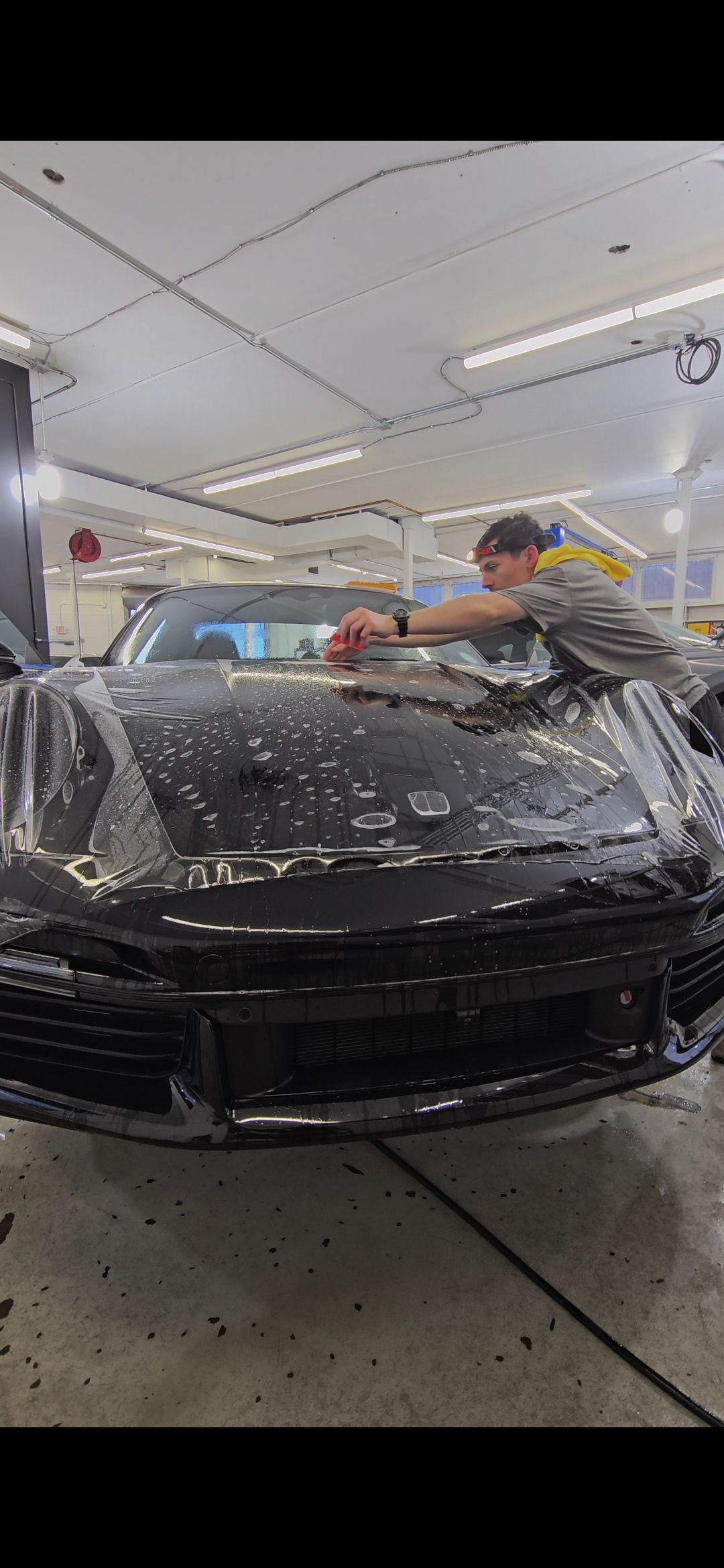 A person working on a black car in a garage, possibly applying a film to it.