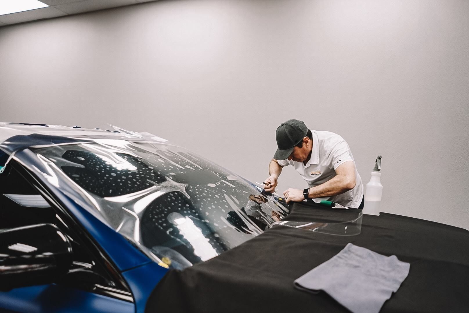 Man applying tint film to a blue car windshield in a workshop.