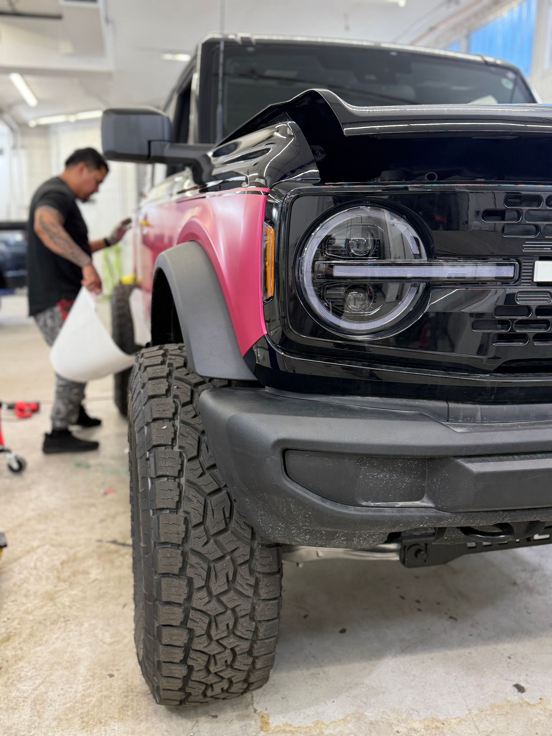 Man wrapping a pink vehicle in a shop. Black Bronco's front tire and light are in focus.