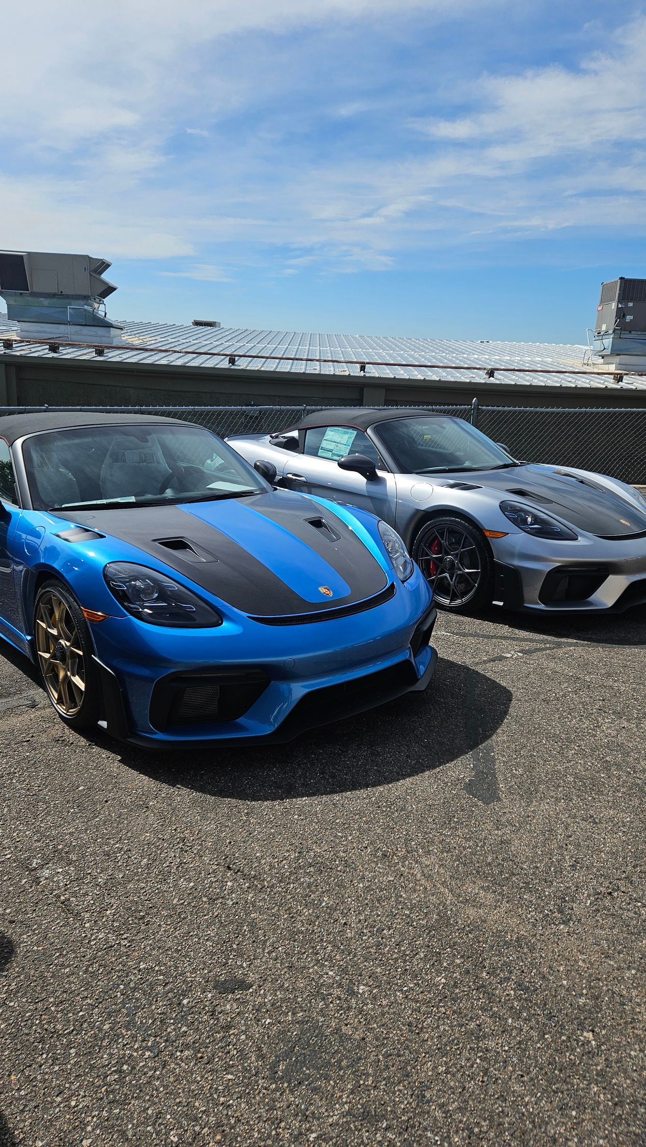 Two Porsche sports cars, one blue with black stripes, and one silver, parked outdoors under a blue sky.