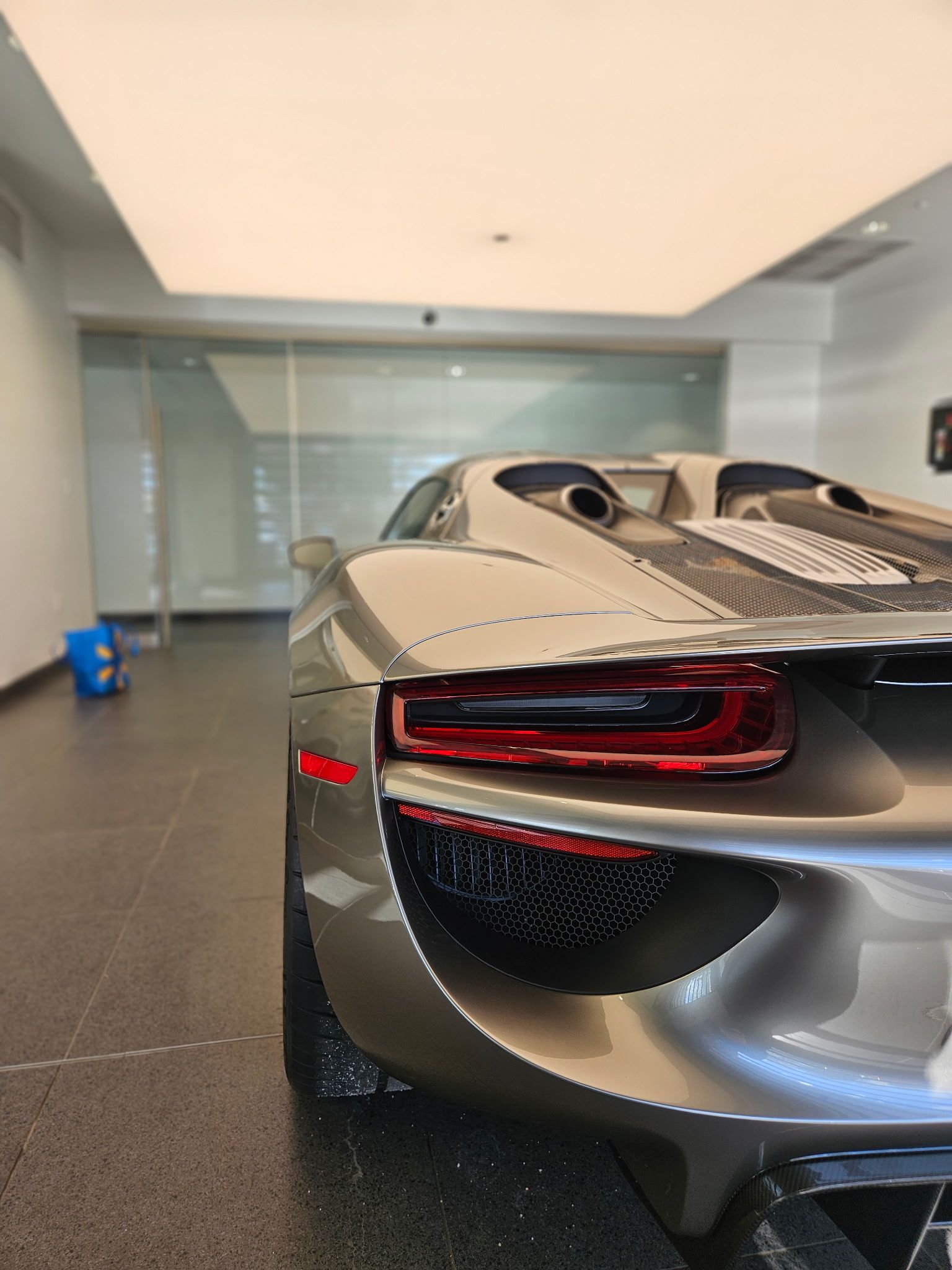 Rear view of a silver Porsche 918 Spyder sports car parked indoors with a red taillight and black grill.