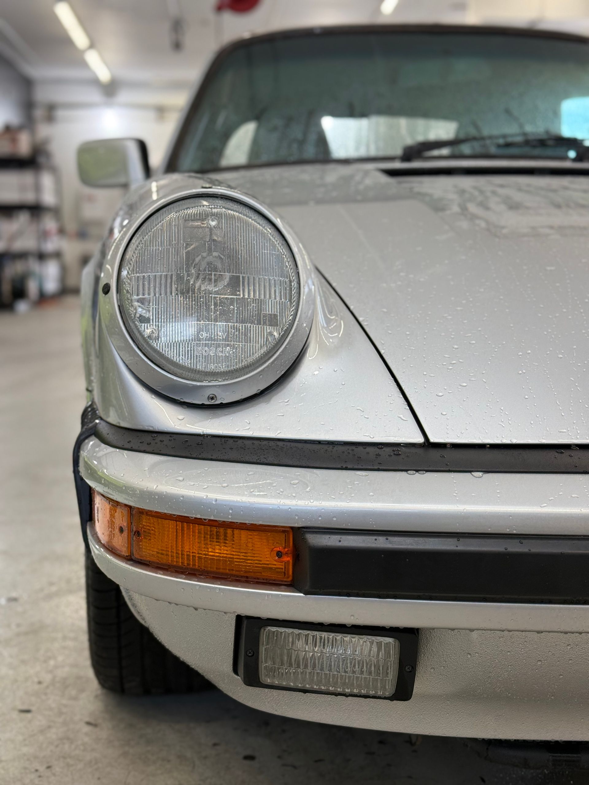 Silver Porsche 911 front, showing headlight, signal, and bumper. Indoors, slightly wet.