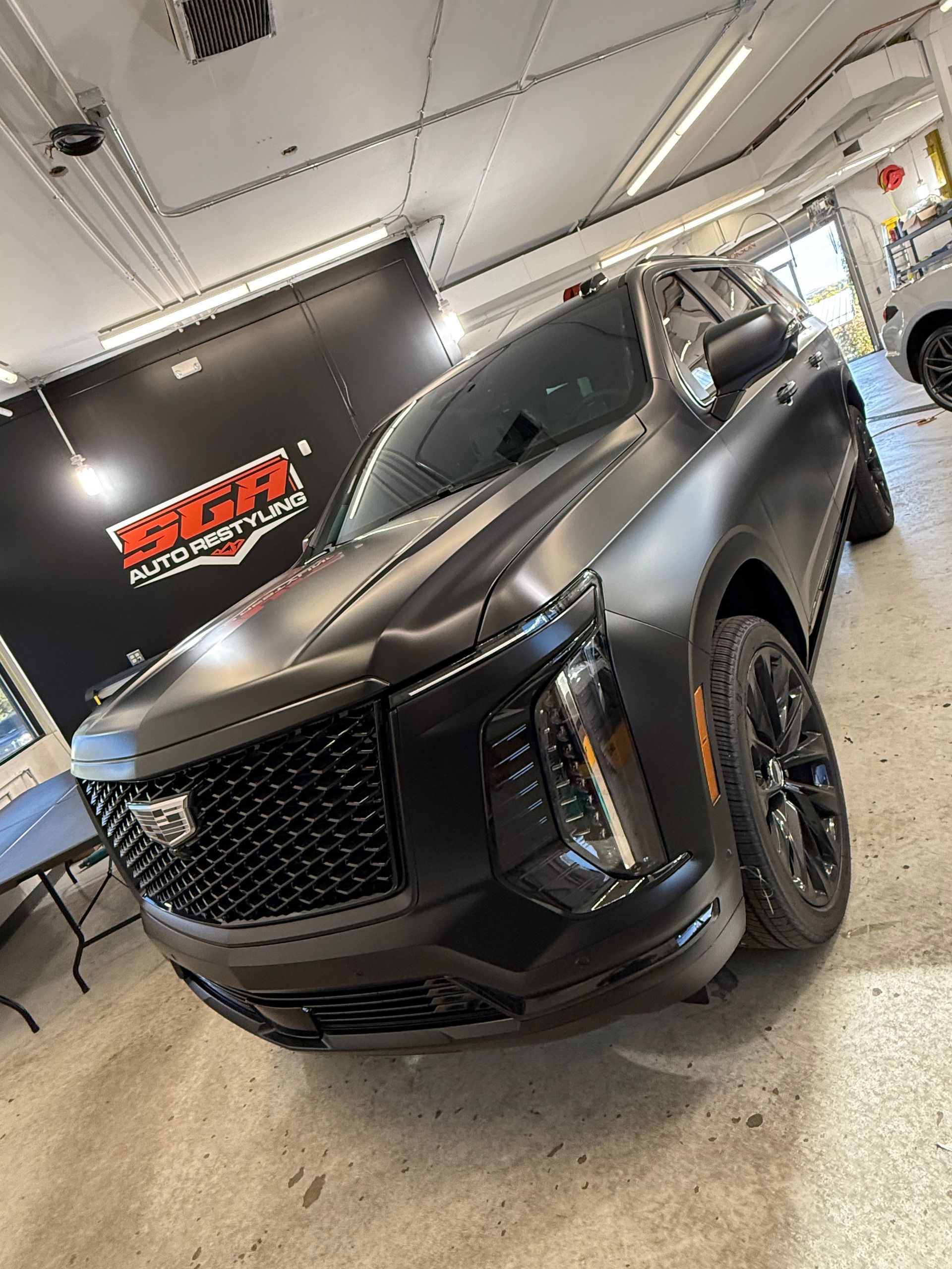 Black Cadillac SUV in a shop, matte finish, tinted windows, black grille.