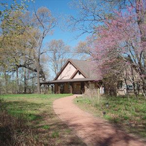 House with porch along a winding dirt path in a grassy yard, framed by trees in spring bloom