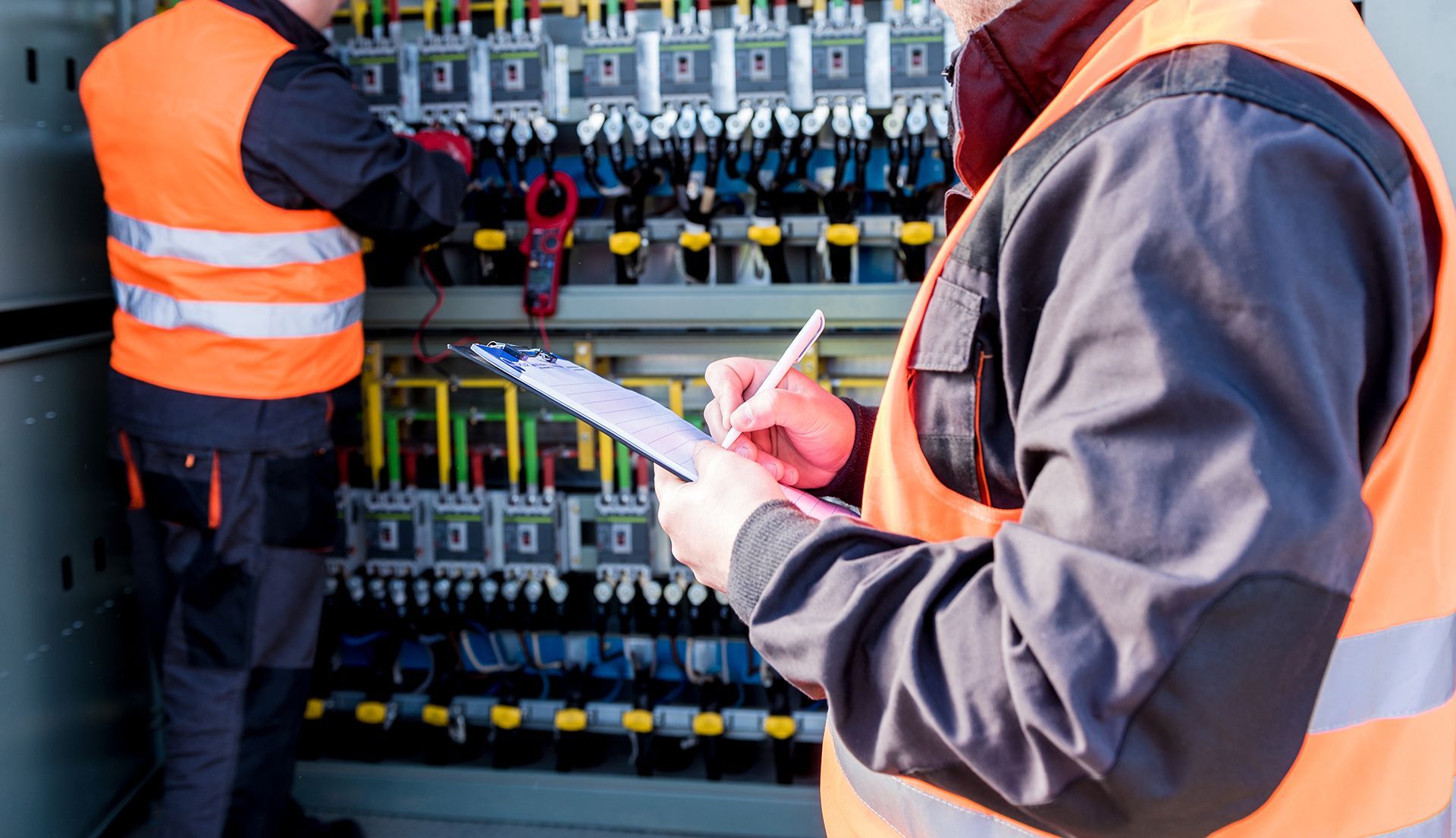 Two men are working on a electrical panel and one is writing on a clipboard