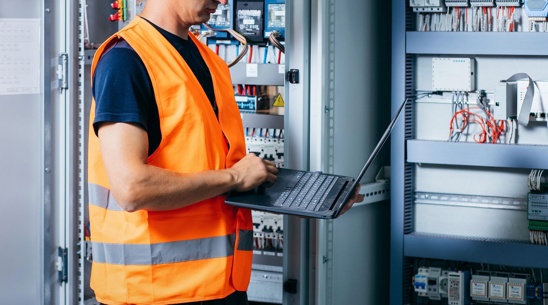 A man in an orange vest is using a laptop computer in an electrical room.