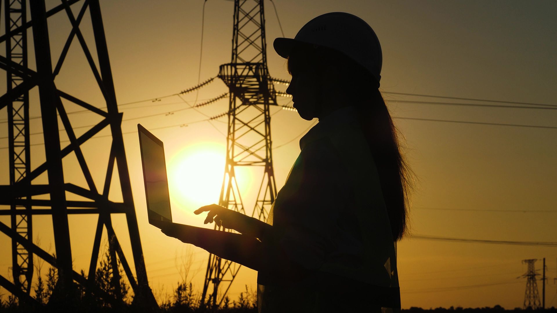A woman is using a laptop computer in front of power lines at sunset.
