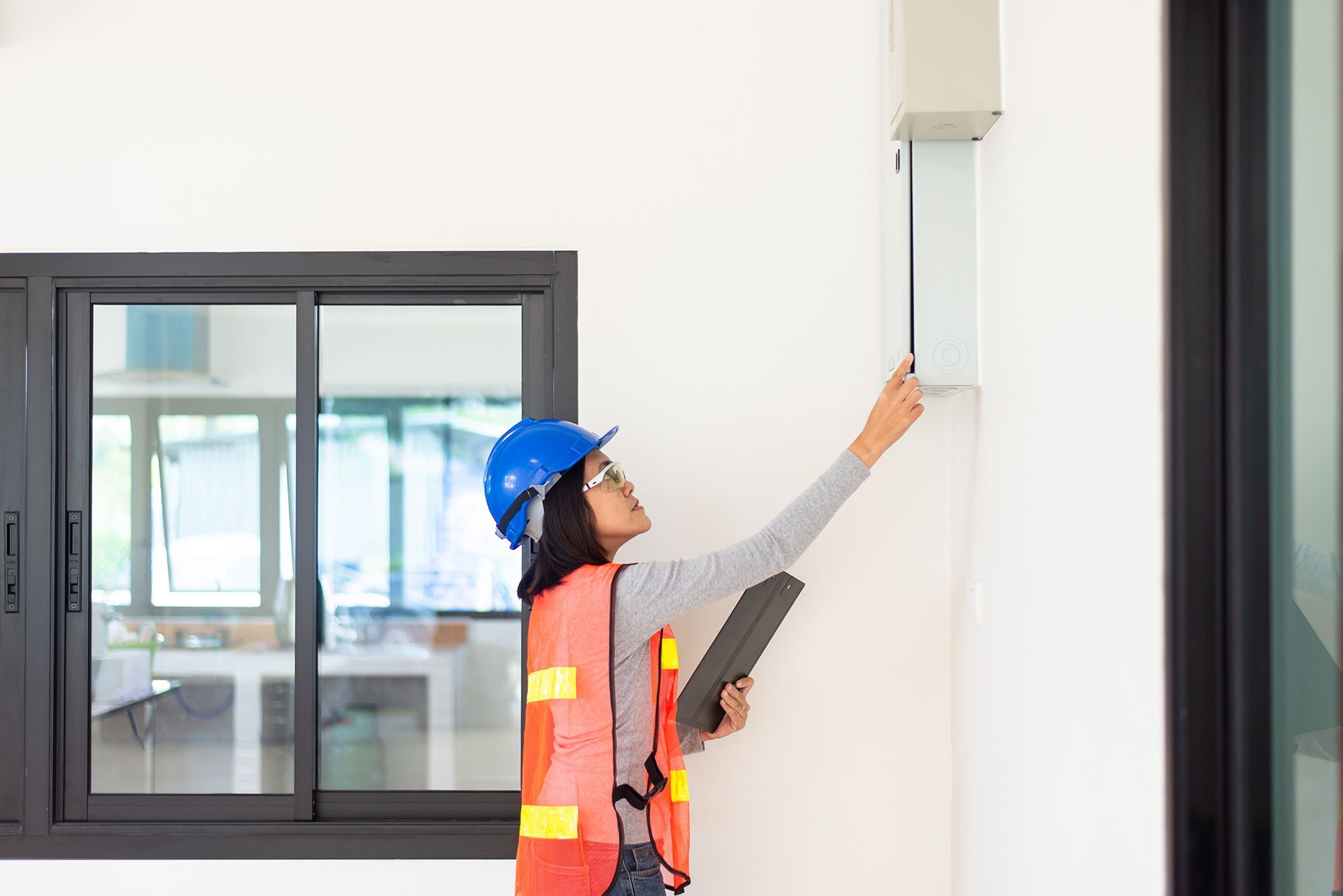 A woman in a hard hat and safety vest is looking at a box on a wall.