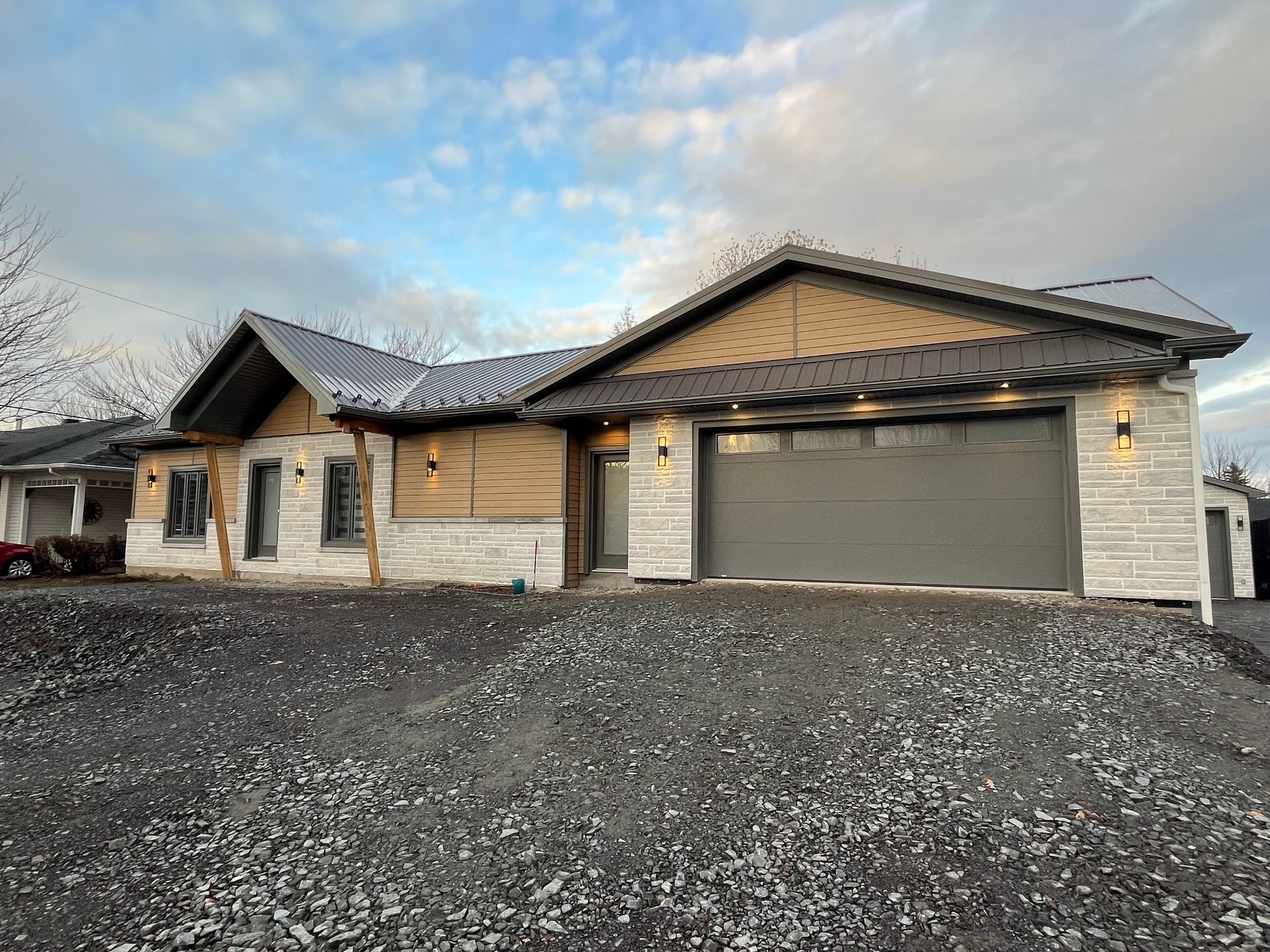 Maison neuve avec porte de garage grise, revêtement en pierre claire et bois, sous un ciel nuageux.